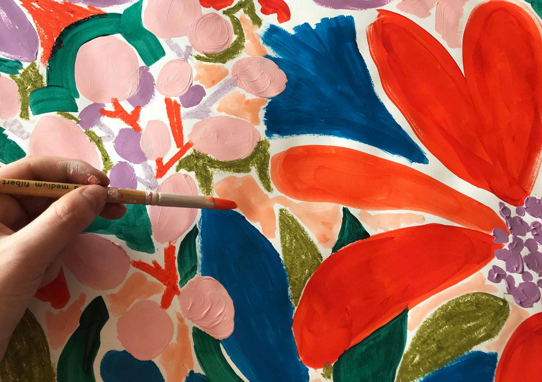 A close-up photograph of a hand painting a colourful floral design on a canvas. The flowers’ colours include orange, blue, pink and green.