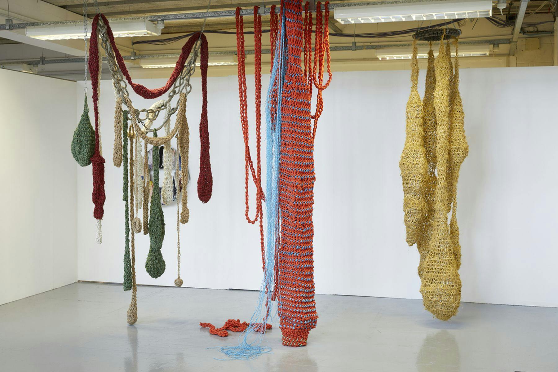 A photograph of three knitted structures hanging from the ceiling. The knitting comprises of greens, oranges, blues, yellows and creams. The structures have varying shapes.