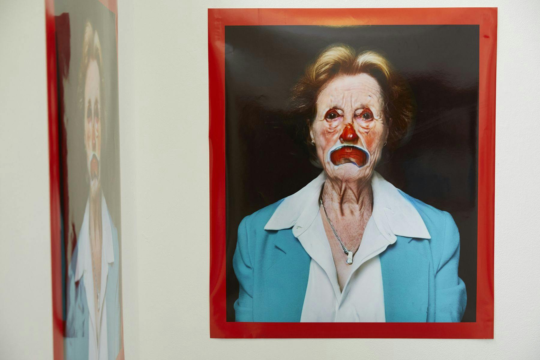 A photograph featuring an image of a person in clown makeup mounted against a white wall. They have a downturned unhappy expression and are wearing a white shirt and blue jacket. The image has a black background and is surrounded by a red frame. The side wall has the same image.