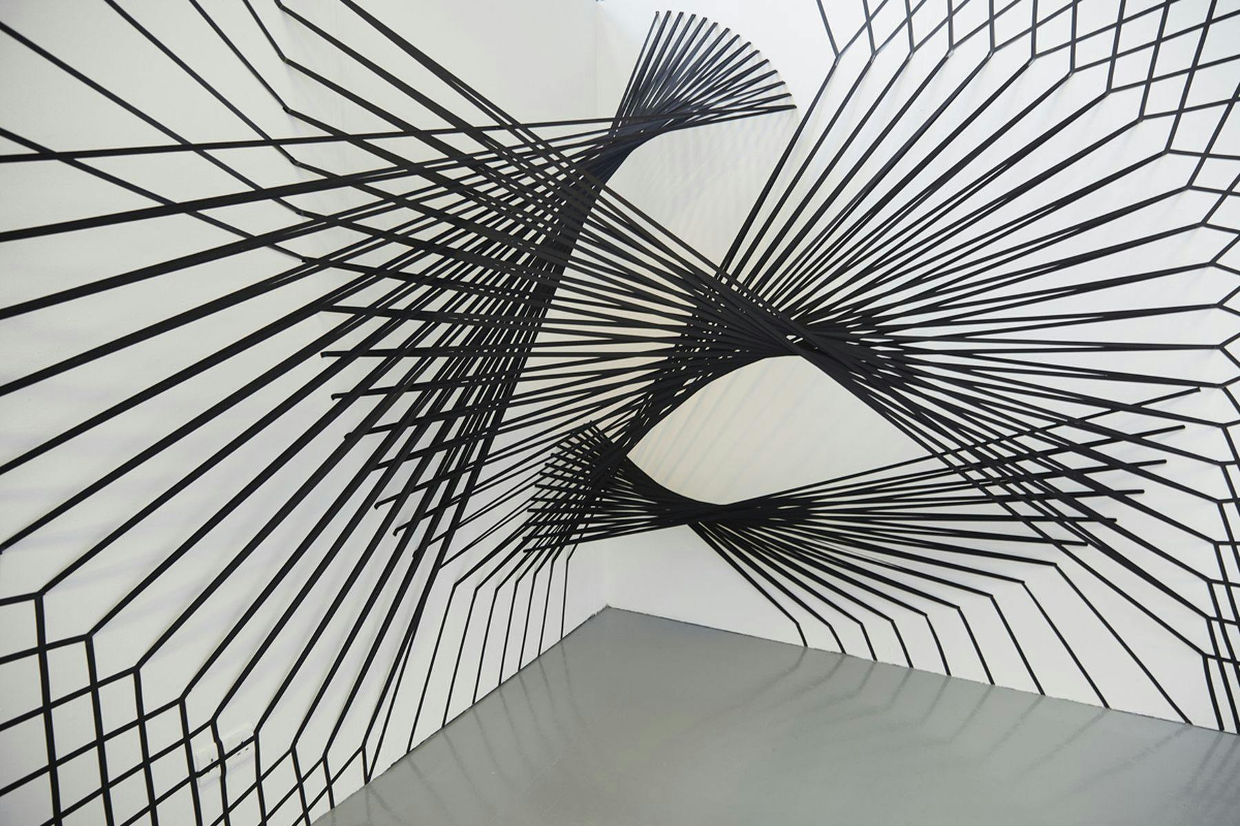 A photograph of black spiral structures mounted against a white wall. There are three interlinking structures in total consisting of vertical lines.