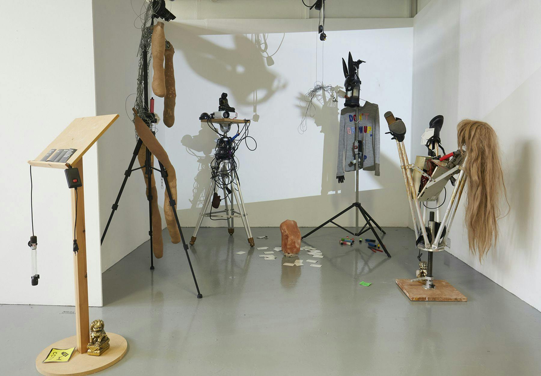 A photograph of an installation featuring a lectern, two tripods and two projector stands, one with a projector on top. They are lined in a row and adorned with different objects including a wig, jumper and small statues. One of the projector stands is upturned. On the floor there are cards, felt tips and crystal.