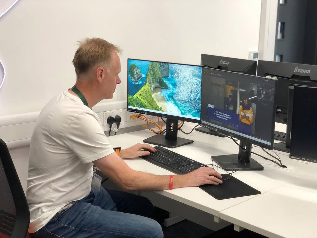 A person in a white shirt works at a desk with two monitors displaying a tropical scene and a graphical interface.