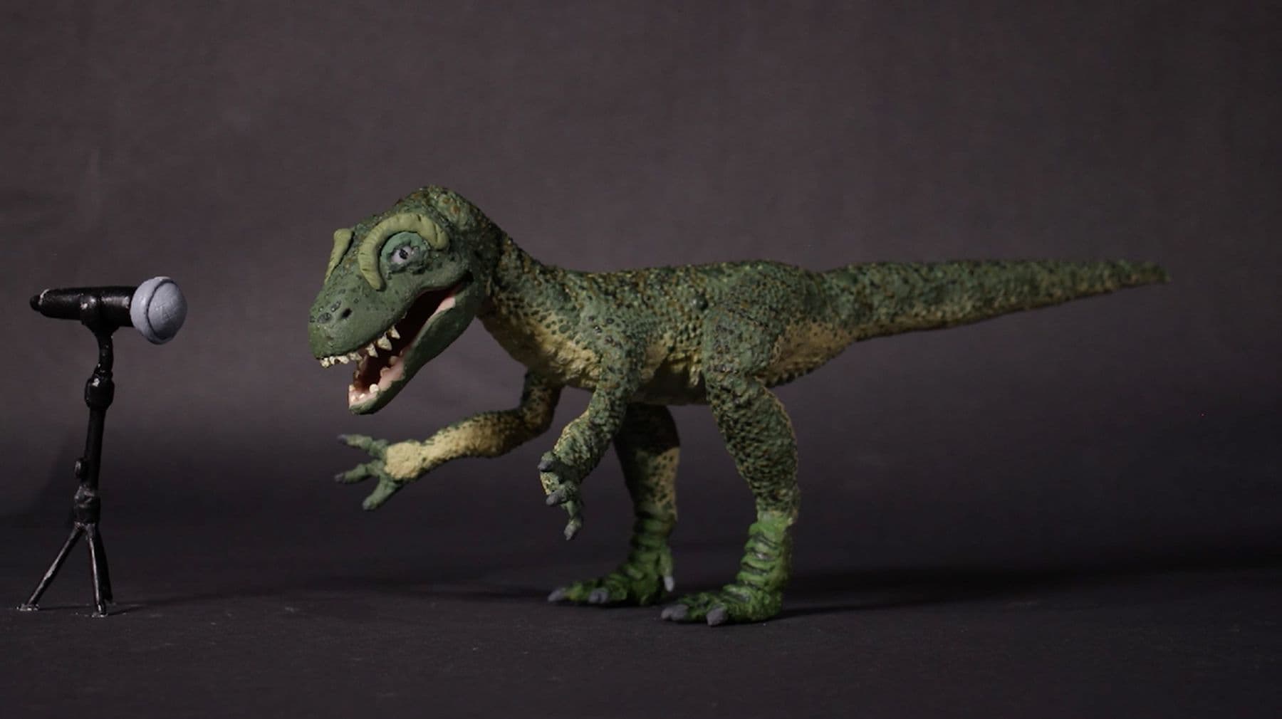 A stop motion still of a plasticine T. rex dinosaur, standing in front of a microphone.