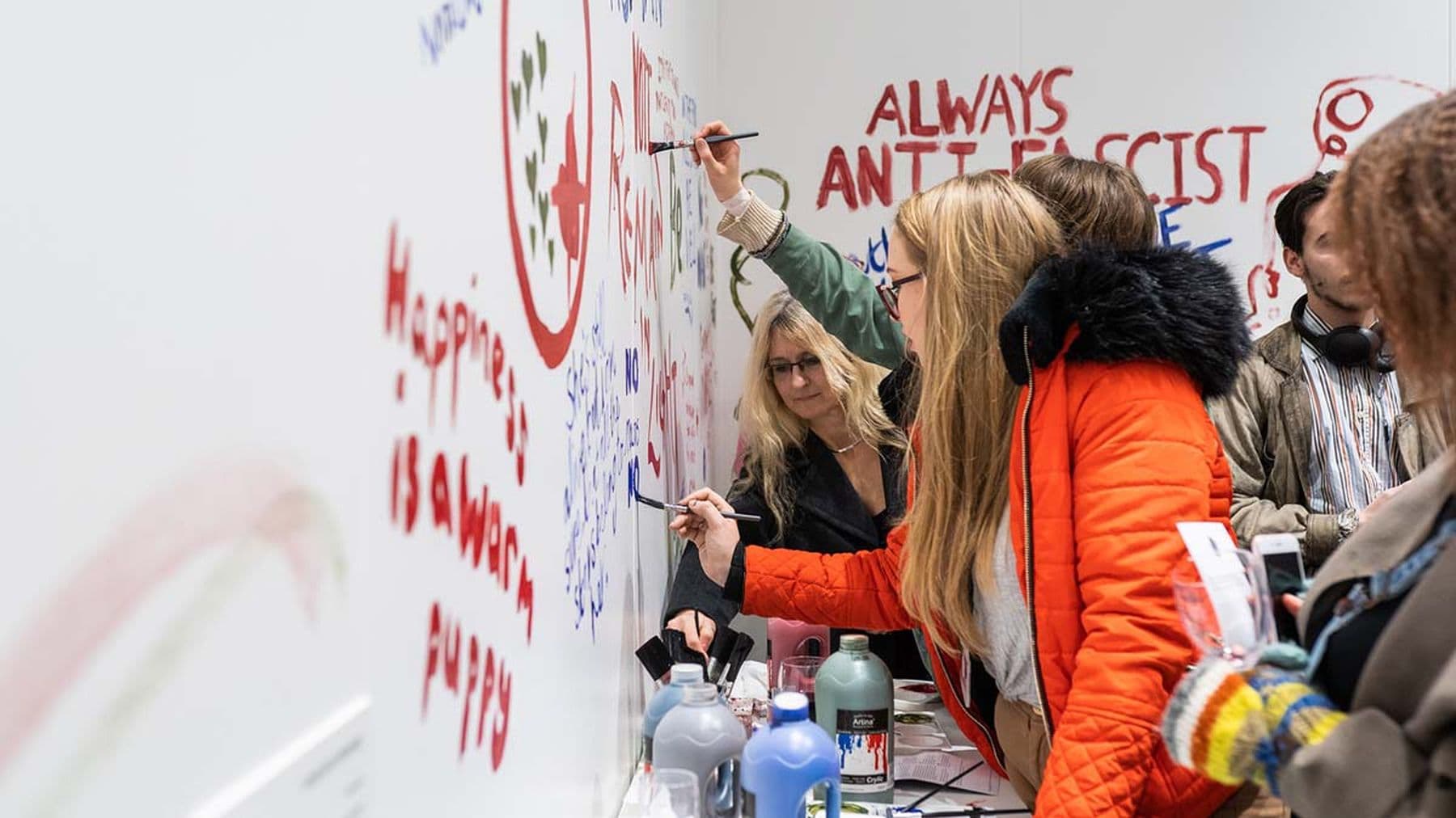 A photograph of people painting and writing messages on a white wall; a woman in an orange coat paints near words that read: 'Always anti facist.'