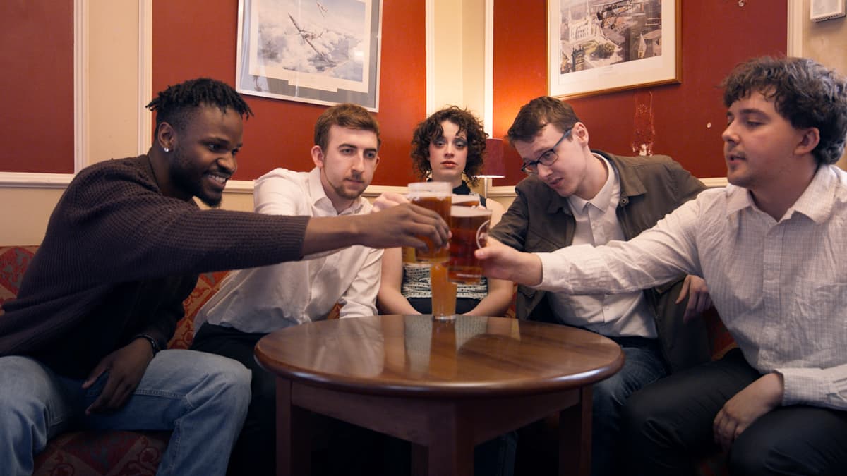 Five people sitting around a table in a pub, clinking glasses in a toast apart from one person in the middle, who does not participate.