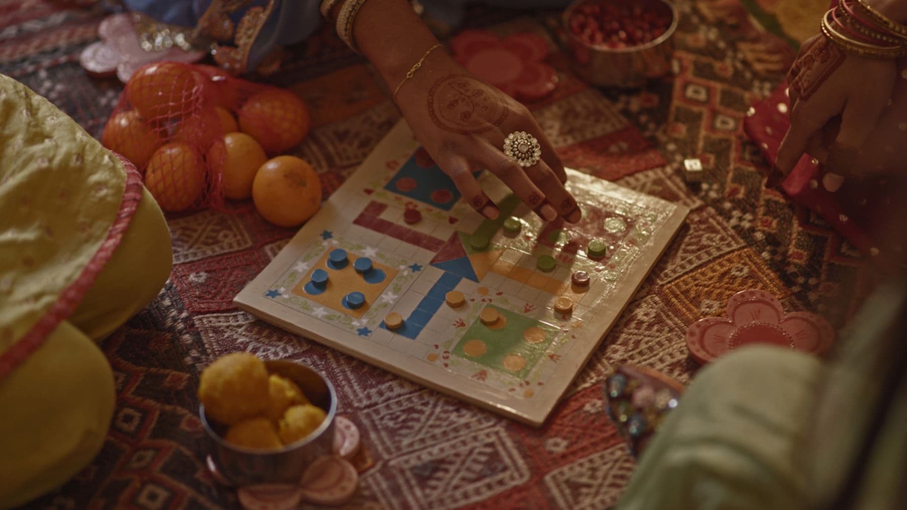 Hands playing a colorful board game on a patterned rug, surrounded by fruits and snacks, with henna designs visible on one hand.
