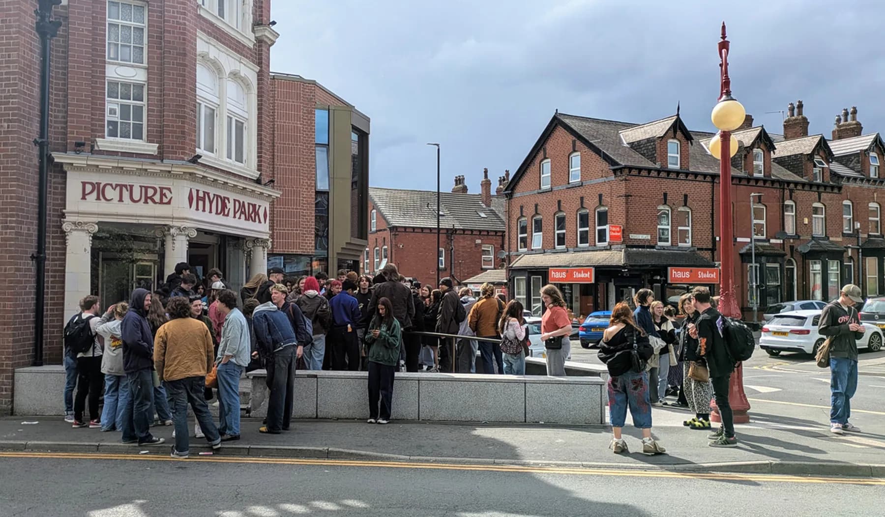 A photograph of a crowd of people gathered outside Hyde Park Picture House, a historic red-brick cinema, with nearby brick buildings and parked cars on a cloudy day.