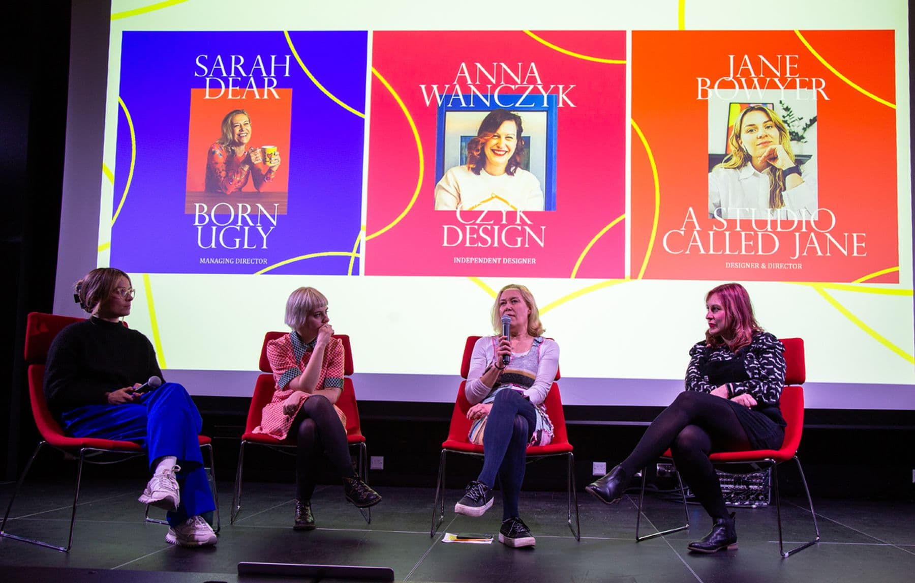 Four women are seated on a stage participating in a panel discussion. A large screen behind displays colorful graphics and names.