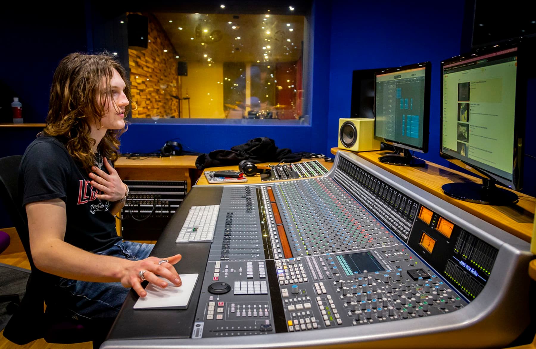 A photograph of a student sittingat a large channel mixing console. They are looking at the computer screen with one hand placed on their chest and another on a touchpad.