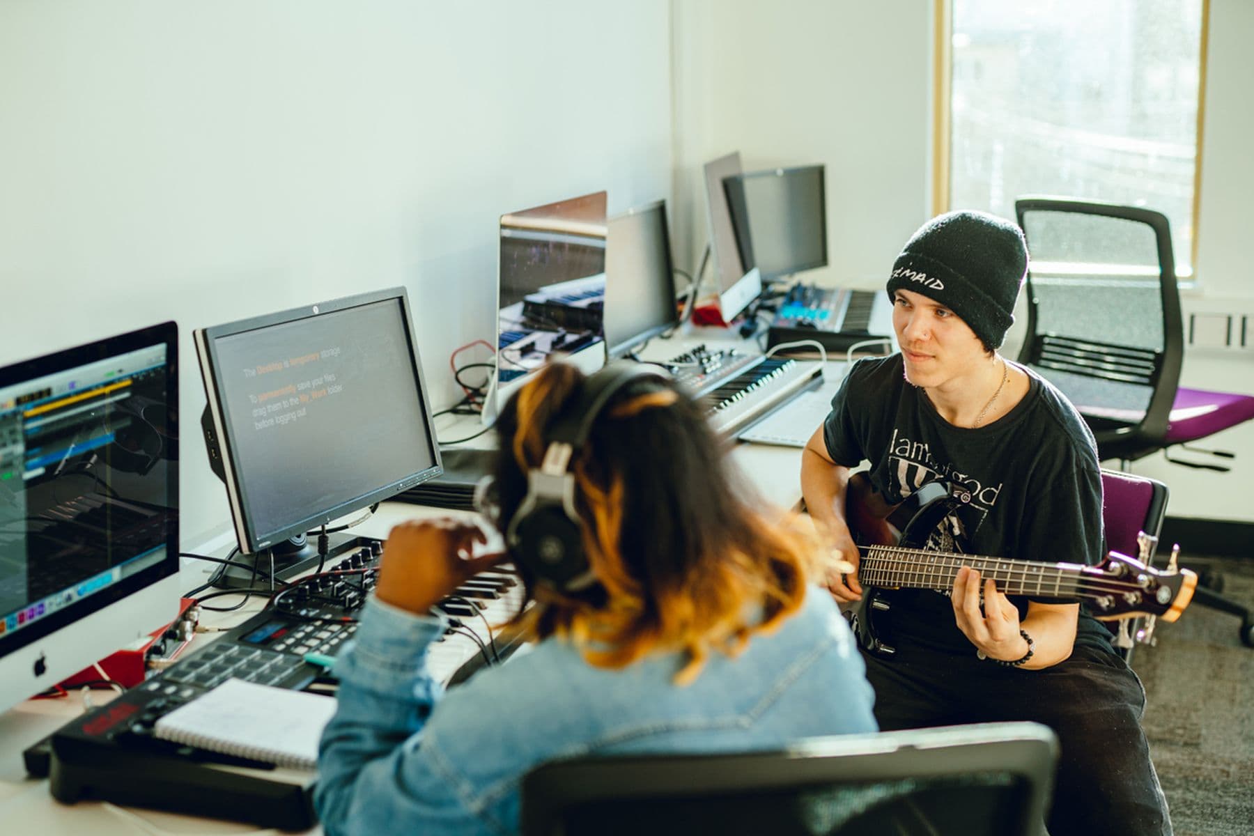 A photograph of two students collaborating together in a classroom. The first student is wearing headphones while looking at a computer screen, the second student is playing the guitar.