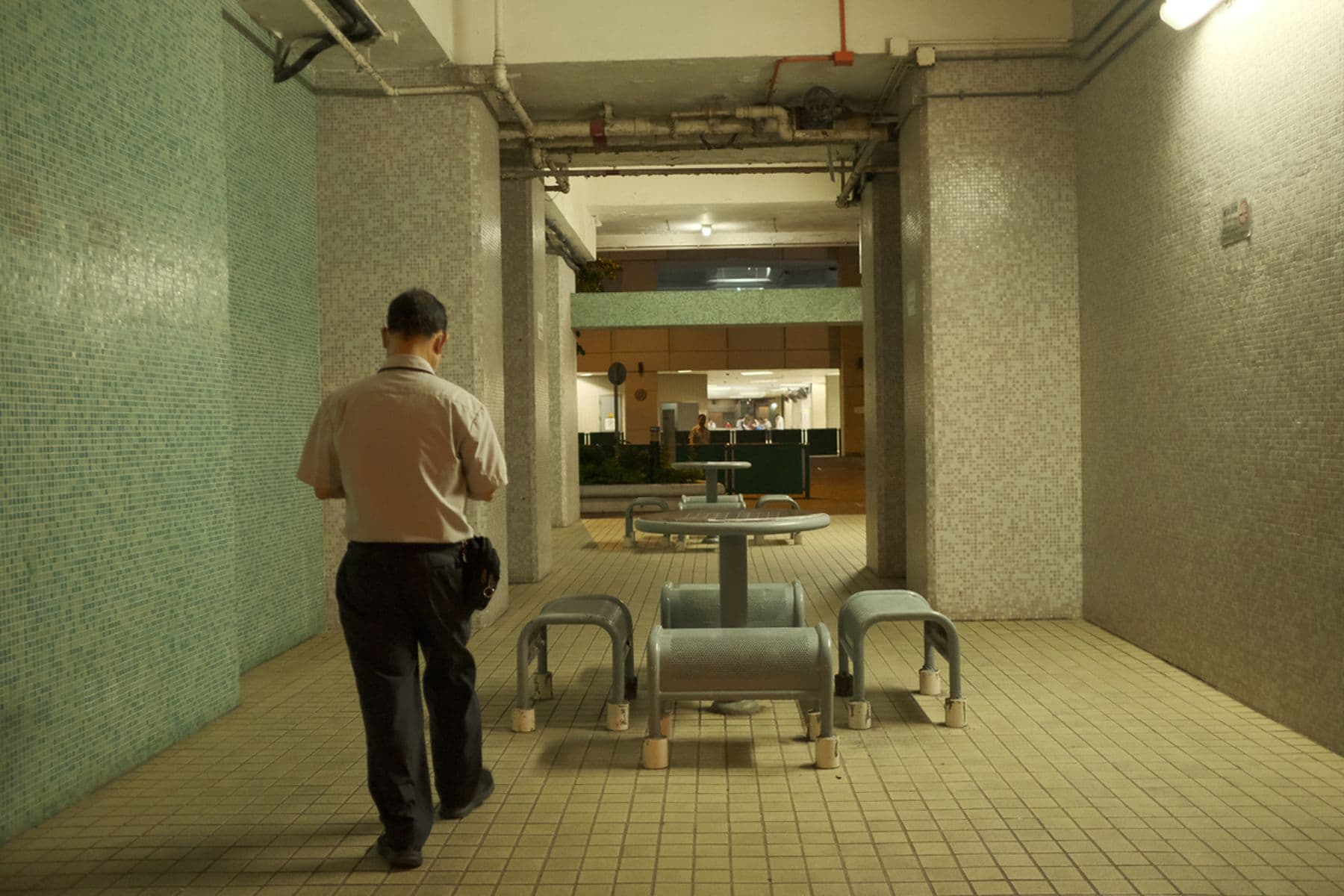 Man walking in an indoor passageway with tile walls, past a metal table and benches, towards a lit area in the distance.