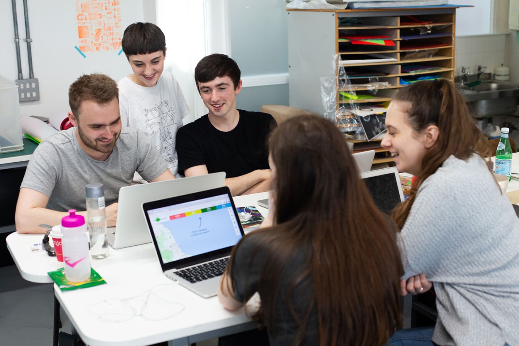 Five people smiling and working together around laptops at a table in a brightly lit studio.