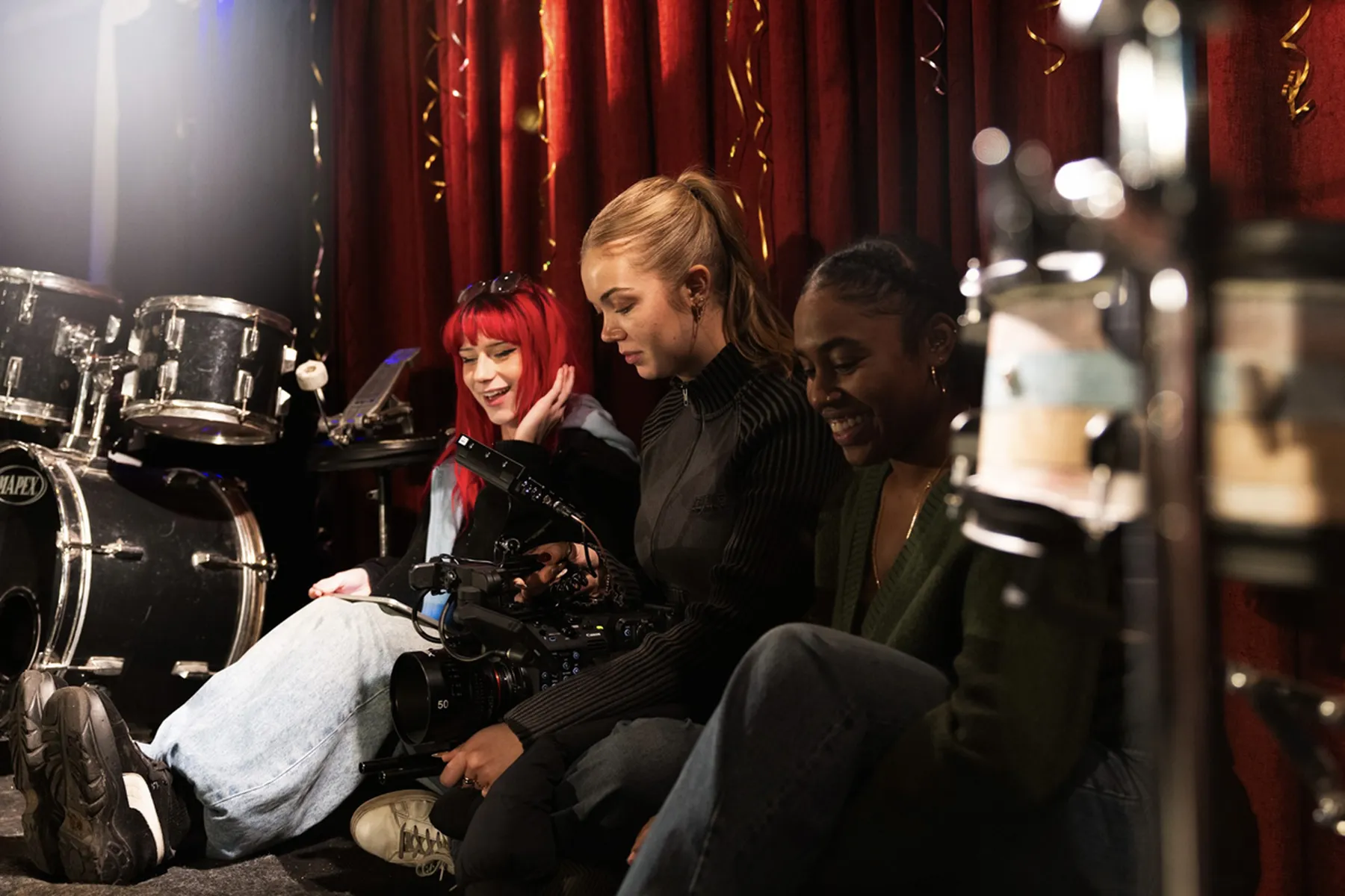 A photograph of three people sitting on a stage floor surrounded by musical instruments looking at a camera with a relaxed and happy demeanour.