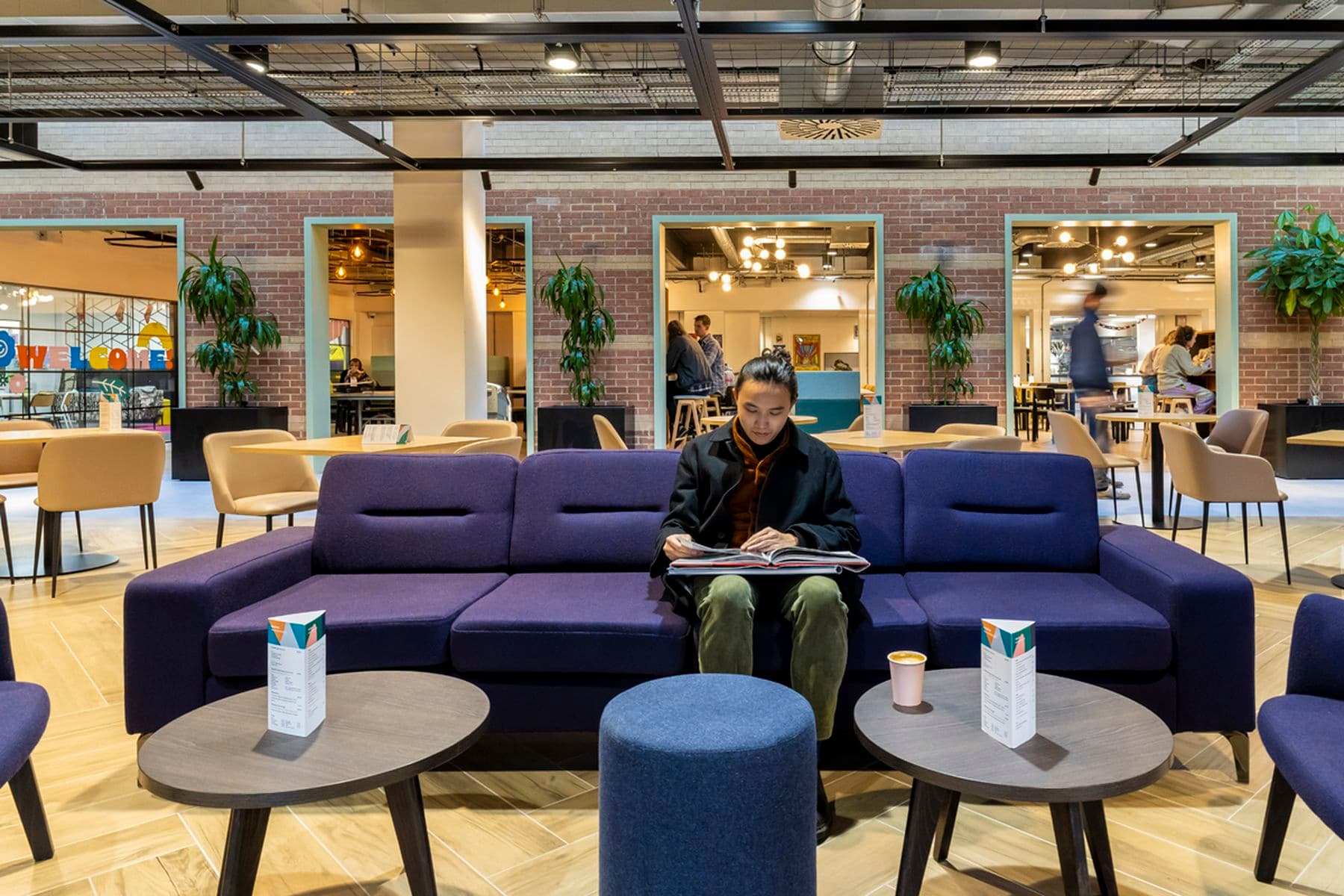 A student sitting on a purple sofa in a modern lounge and cafe area, looking through a book. Tables, chairs, and plants are around; people are in the background.