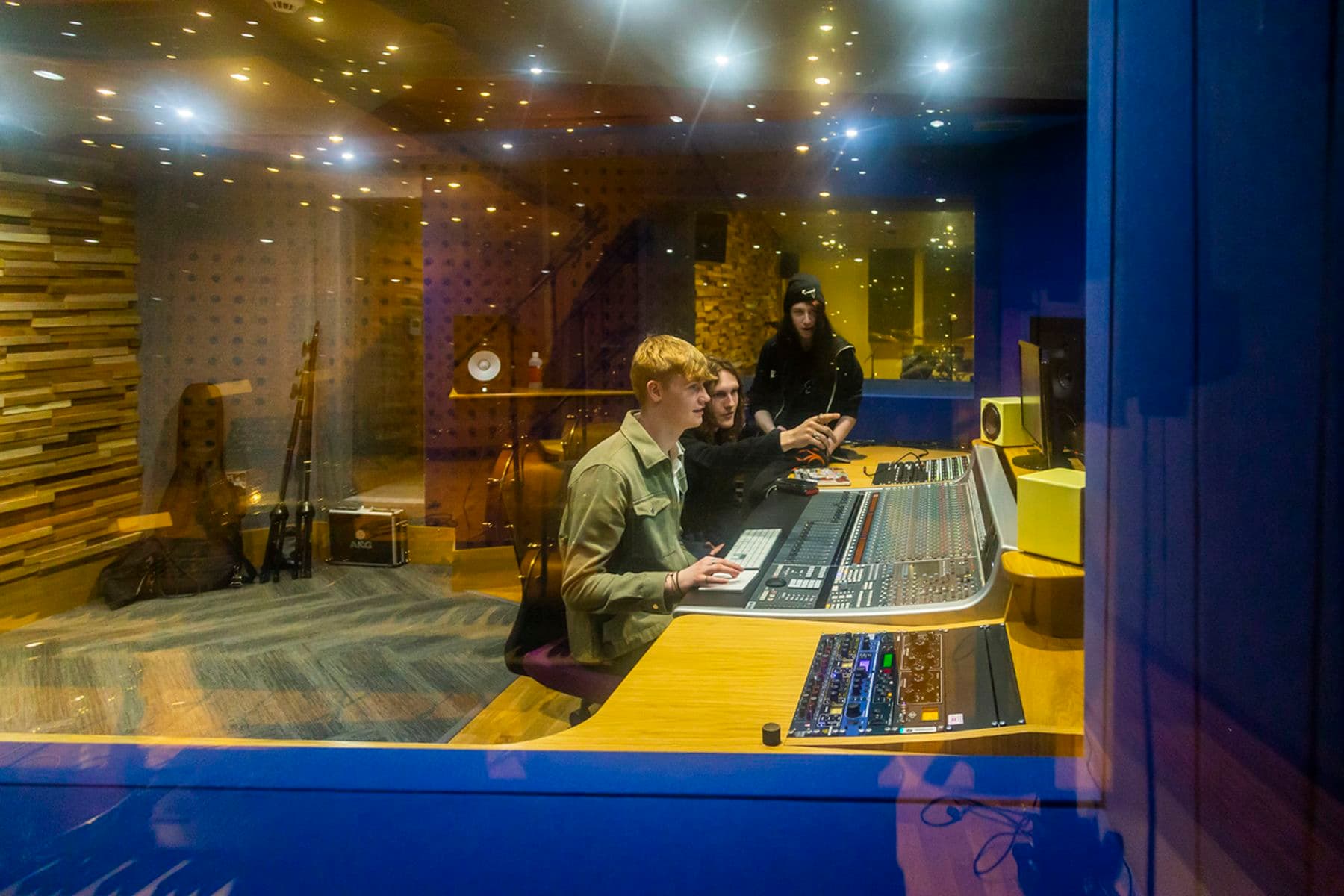 A photograph taken through a recording studio window featuring three students. One of the students points to a computer screen whilst the other looks on. There is another student watching in the background.