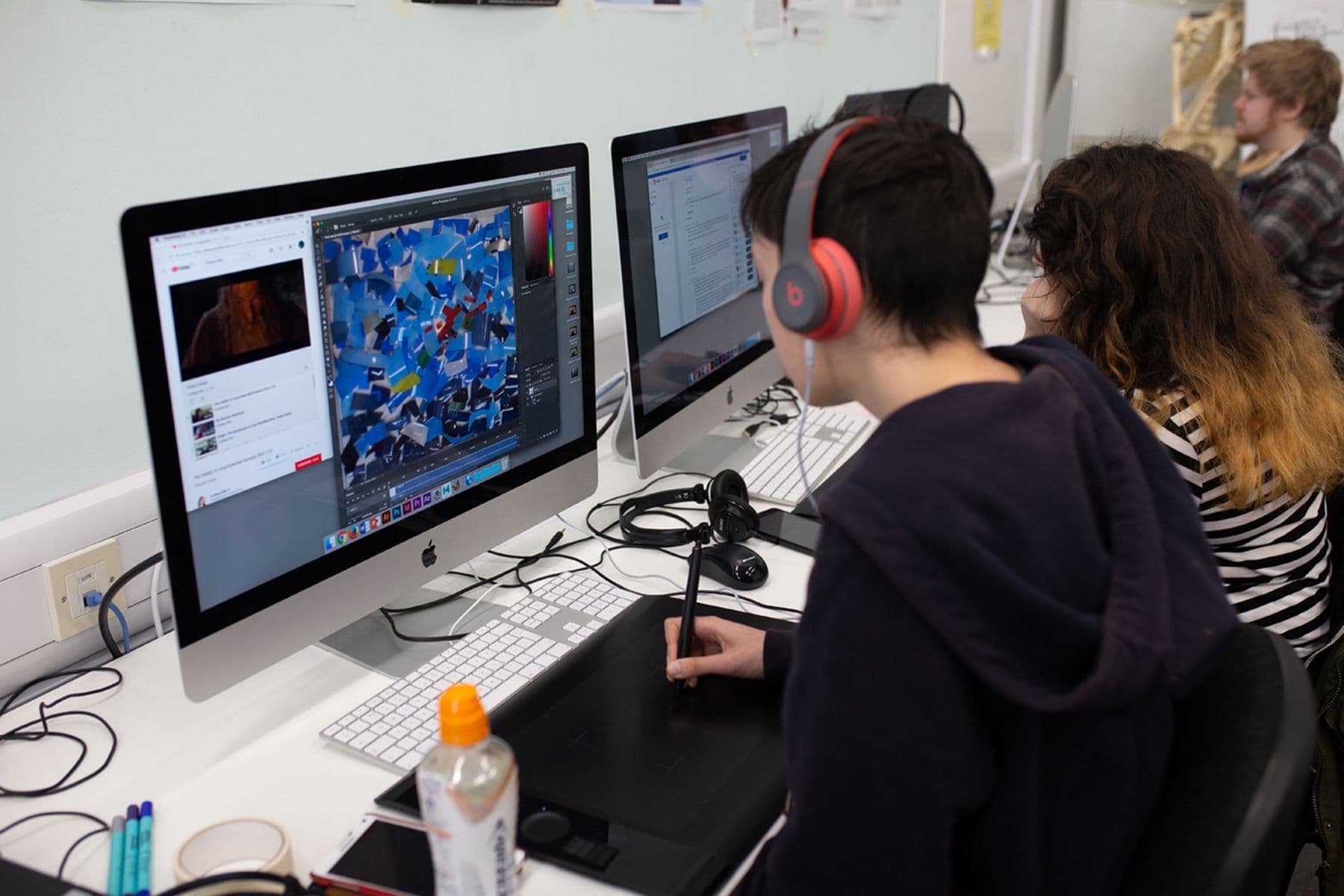 A group of students working on Macs, one using a drawing tablet. Headphones and a drink bottle are visible.