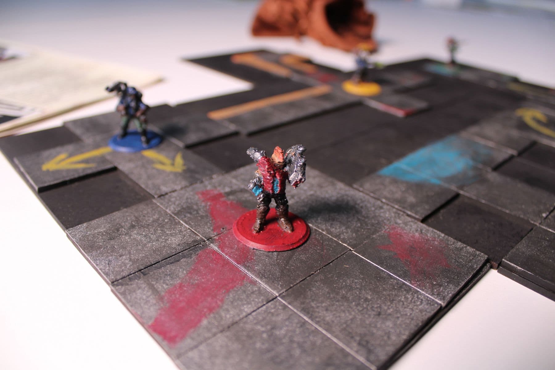 An image featuring miniature figures on a tiled board game map. The red figure stands on a red base whilst the blue figure stands on a blue base.