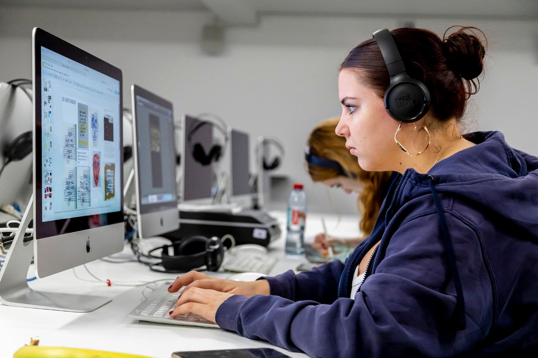 A student wearing headphones works on a computer in a Mac suite, focused on the screen, with another student in the background.