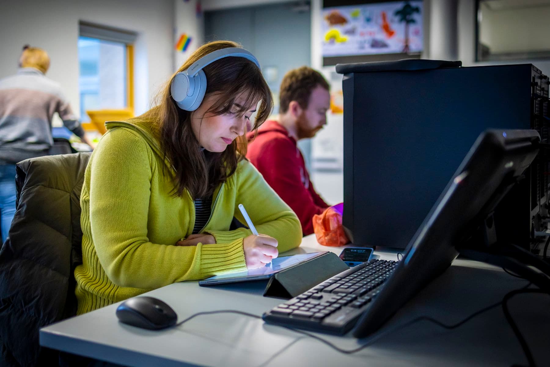 A student in a yellow sweater and headphones writing on a tablet at a desk with a computer, while another person works in the background.