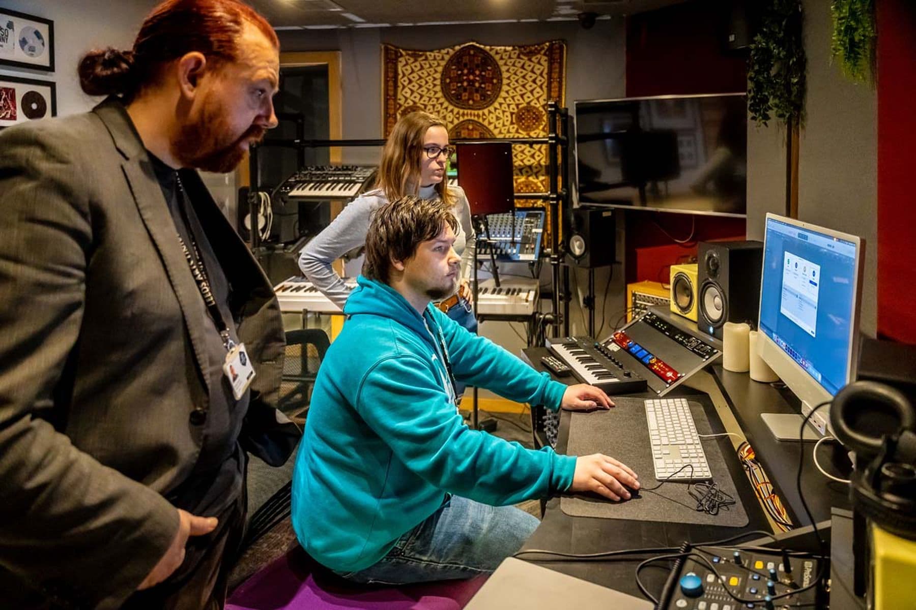 A photograph of three people in a music studio focusing on a computer screen. A person in a blue hoodie operates the computer, surrounded by audio equipment.