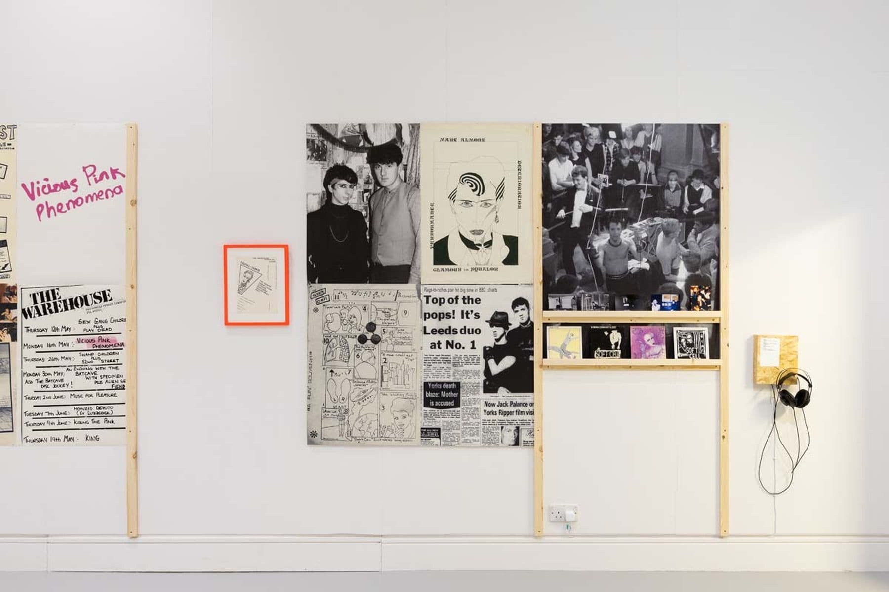 A photograph of an exhibition displaying posters, newspaper clippings, photographs, and music albums, with headphones hanging on the wall beside them.