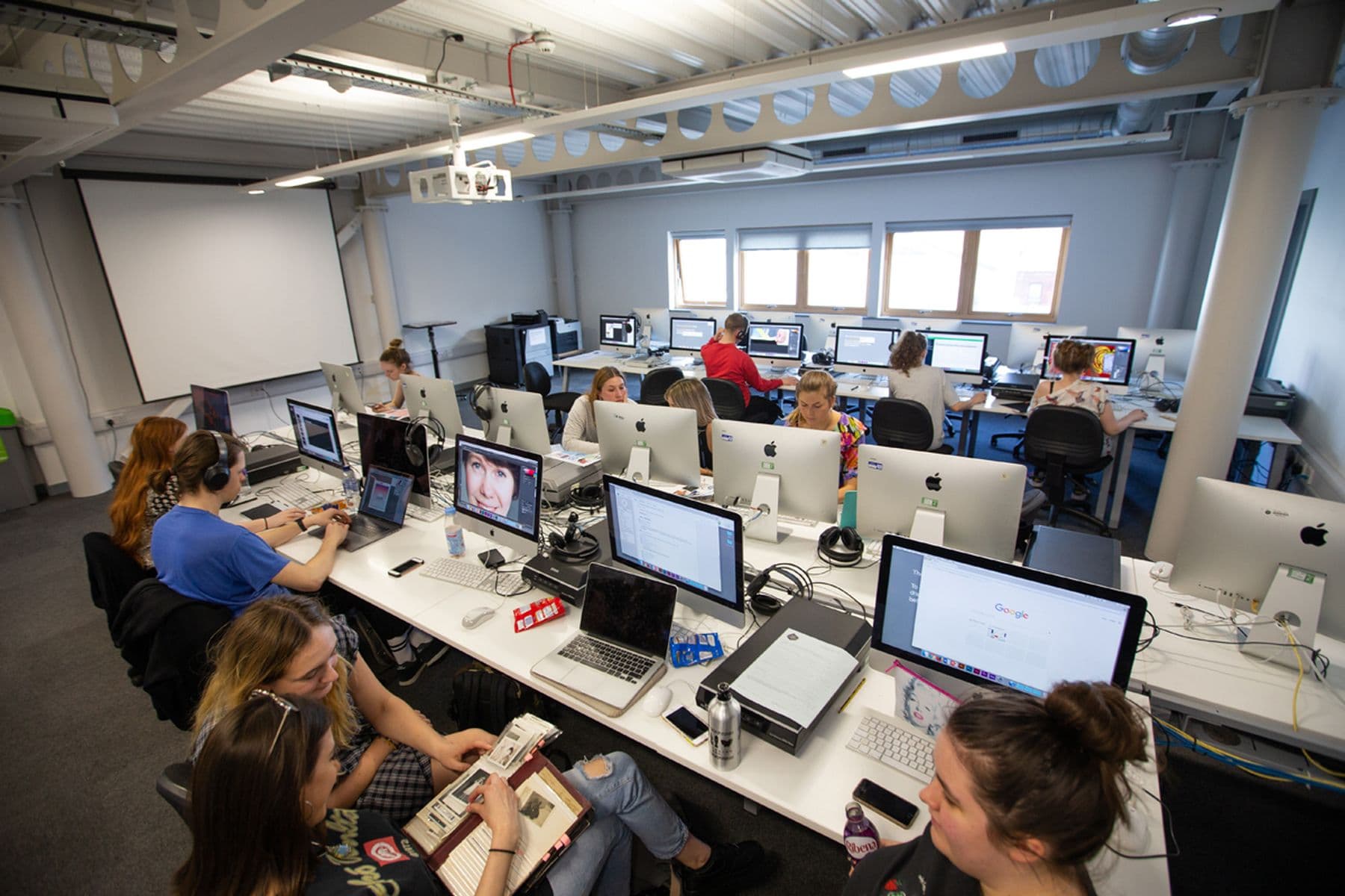 A modern studio with students working on Macs. Some are engaged in conversation, while others focus on their screens.