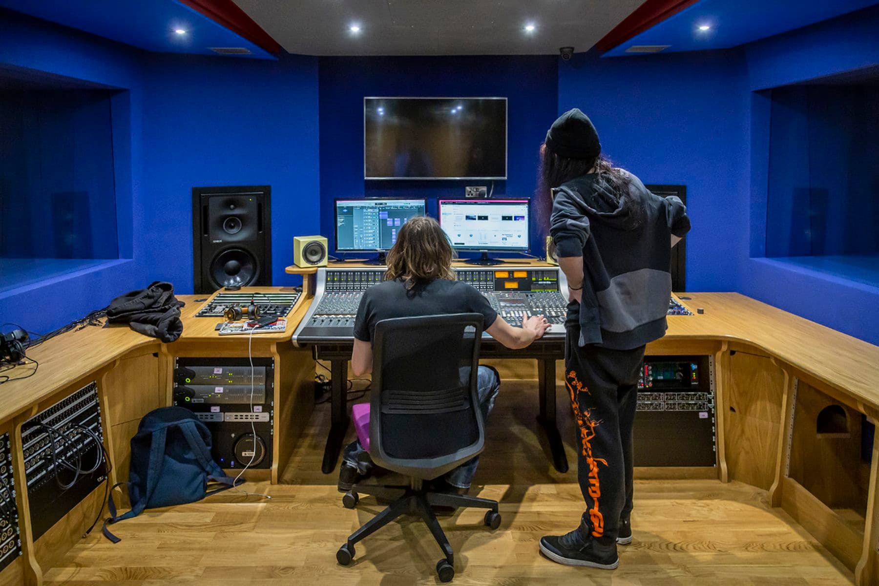 A photograph of two students with their backs turned to the camera in a recording studio. One is sitting down whilst adjusting the dials on a large channel mixing console, the other is standing and watching.