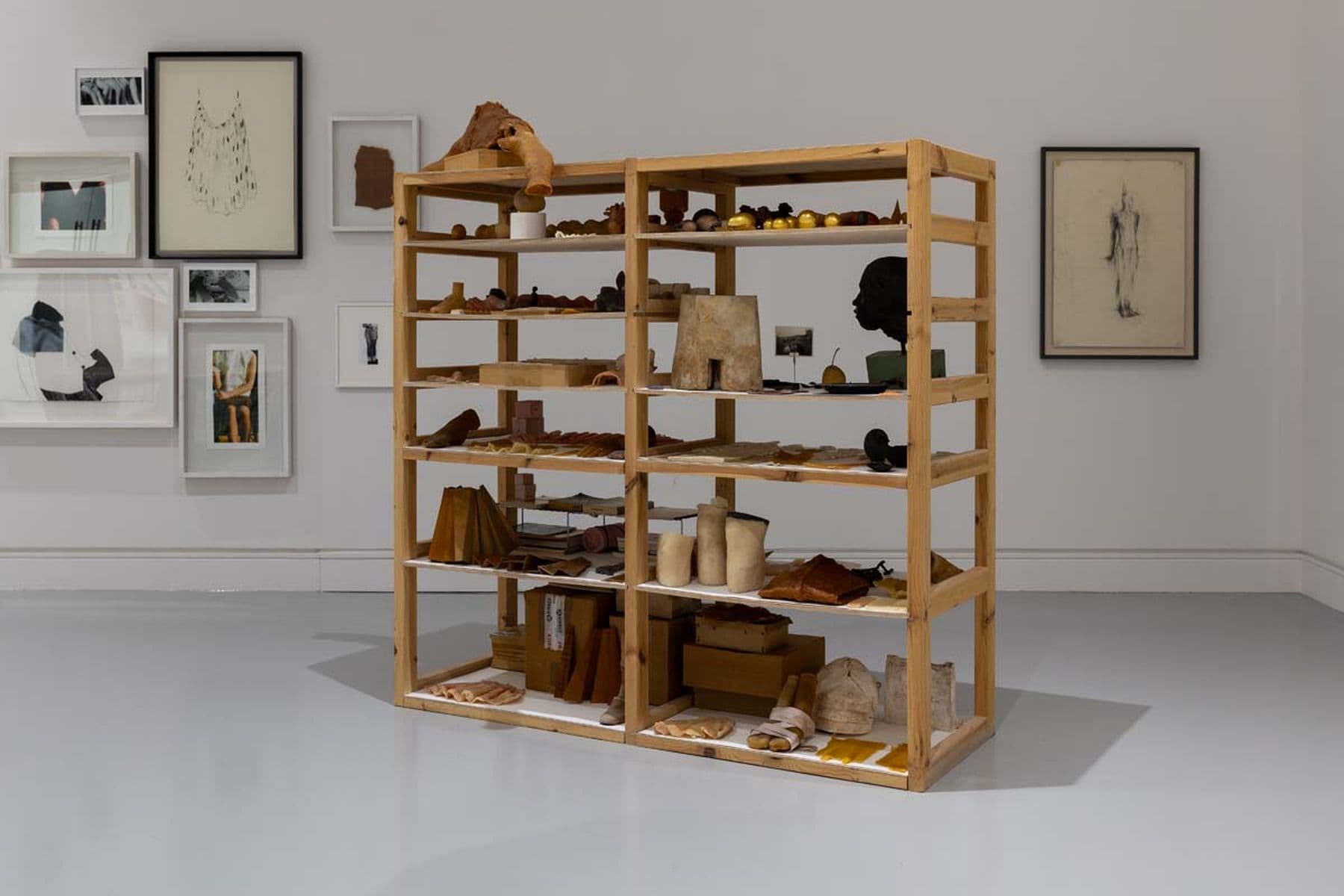 A photograph of a room with wooden shelves displaying various sculptures, with framed prints on the wall in the background.
