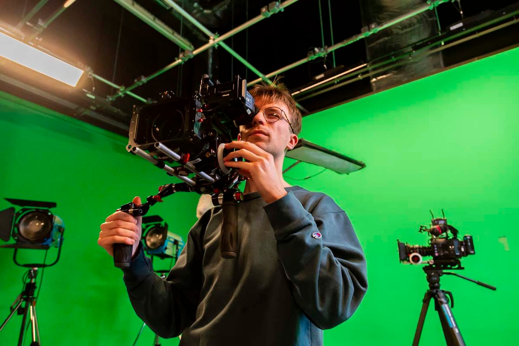 Person holding a professional camera in a green screen studio with lighting and equipment in the background.
