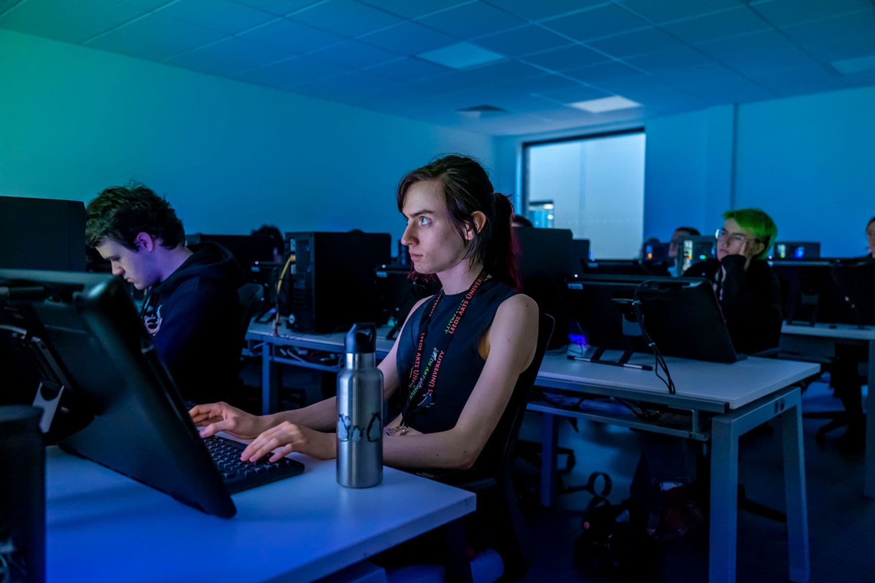 Students working on computers in a dimly lit room, focused on screens. A water bottle is on the desk beside one person.