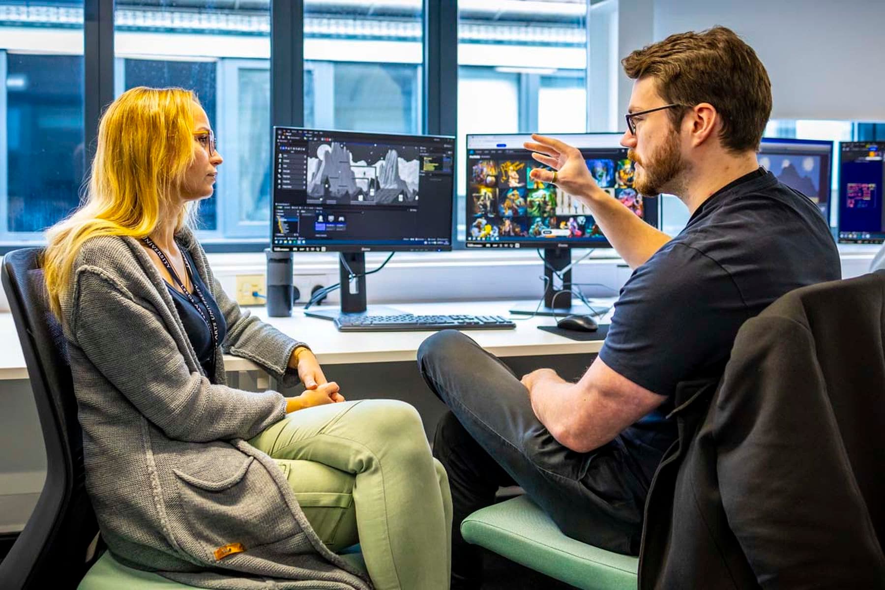 A photograph of two people in a studio having a conversation in front of computer monitors displaying  colourful designs.