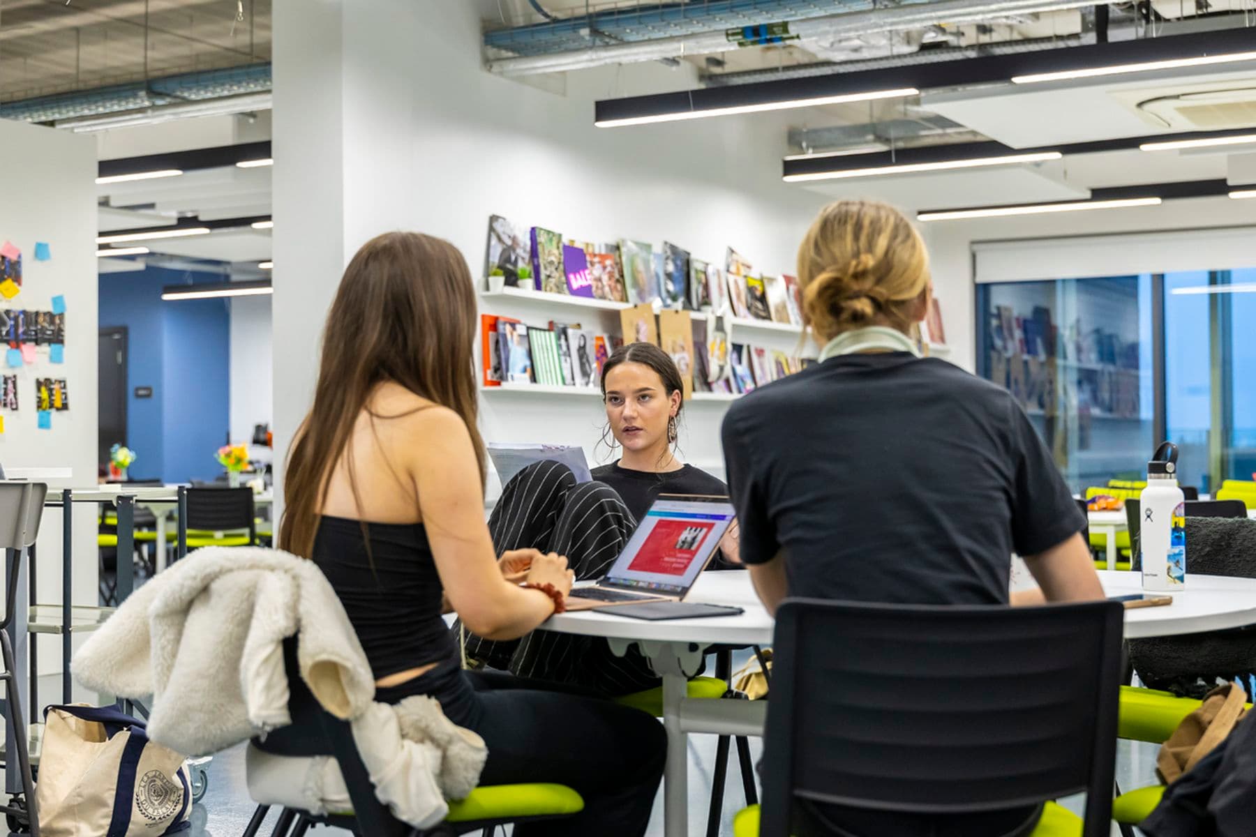 Students having a discussion around a table in a studio, while one uses a laptop. Books and magazines line the wall in the background.