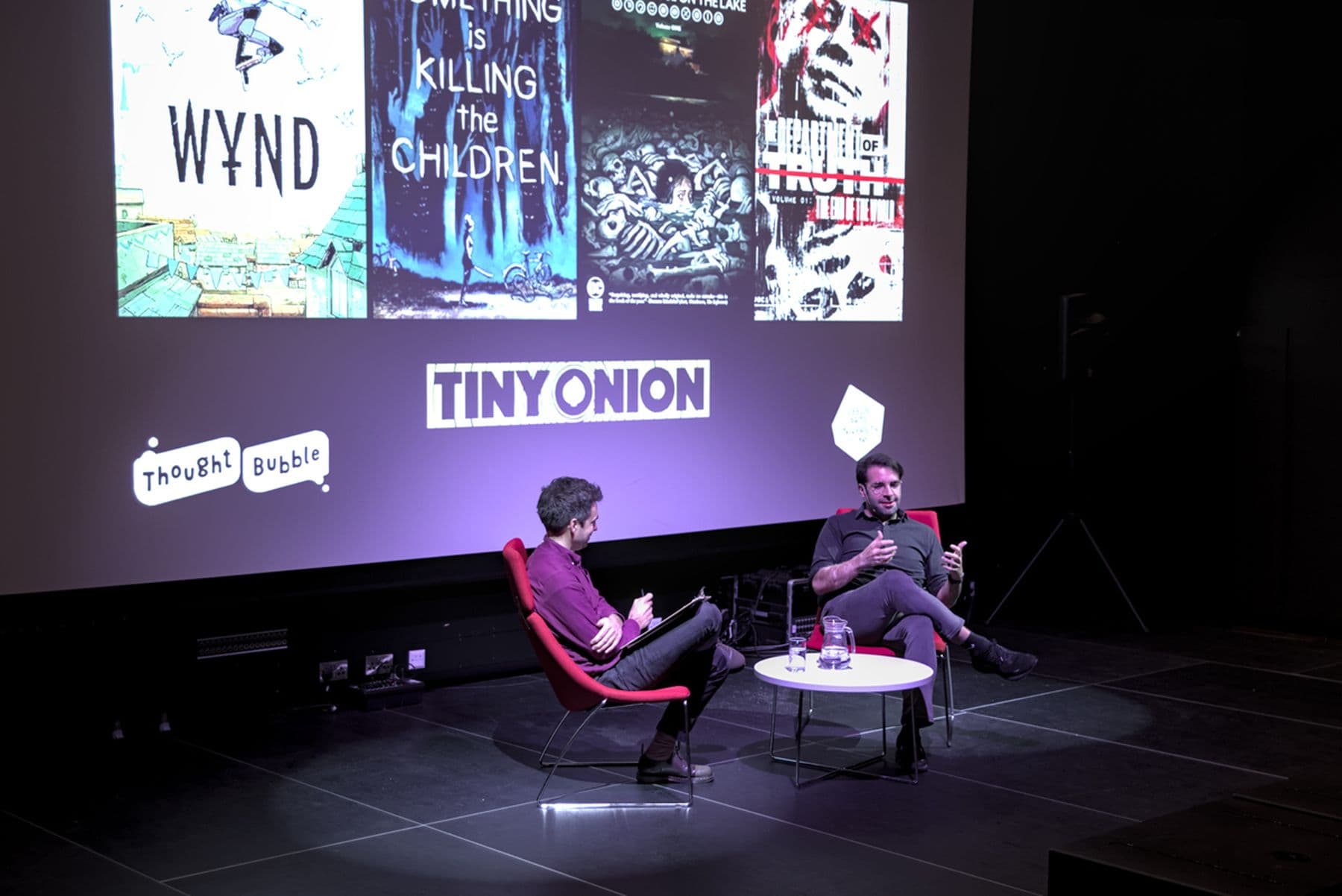 A photograph of two people seated on a stage discussing comics, with a large screen displaying comic book covers and logos in the background.