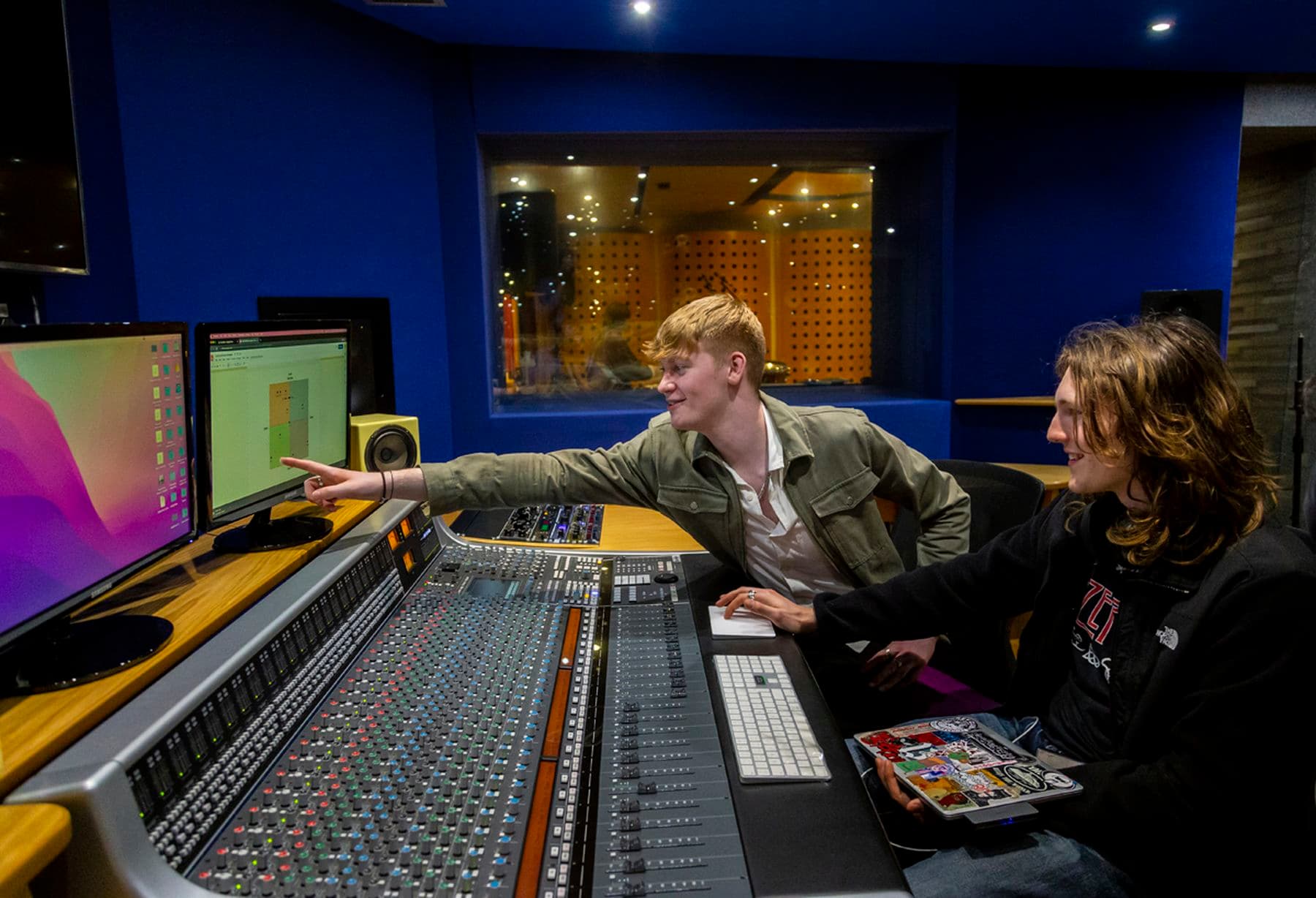 A photograph of two students in a recording studio. One is pointing at the computer screen while the other is holding a laptop and using a touchpad.