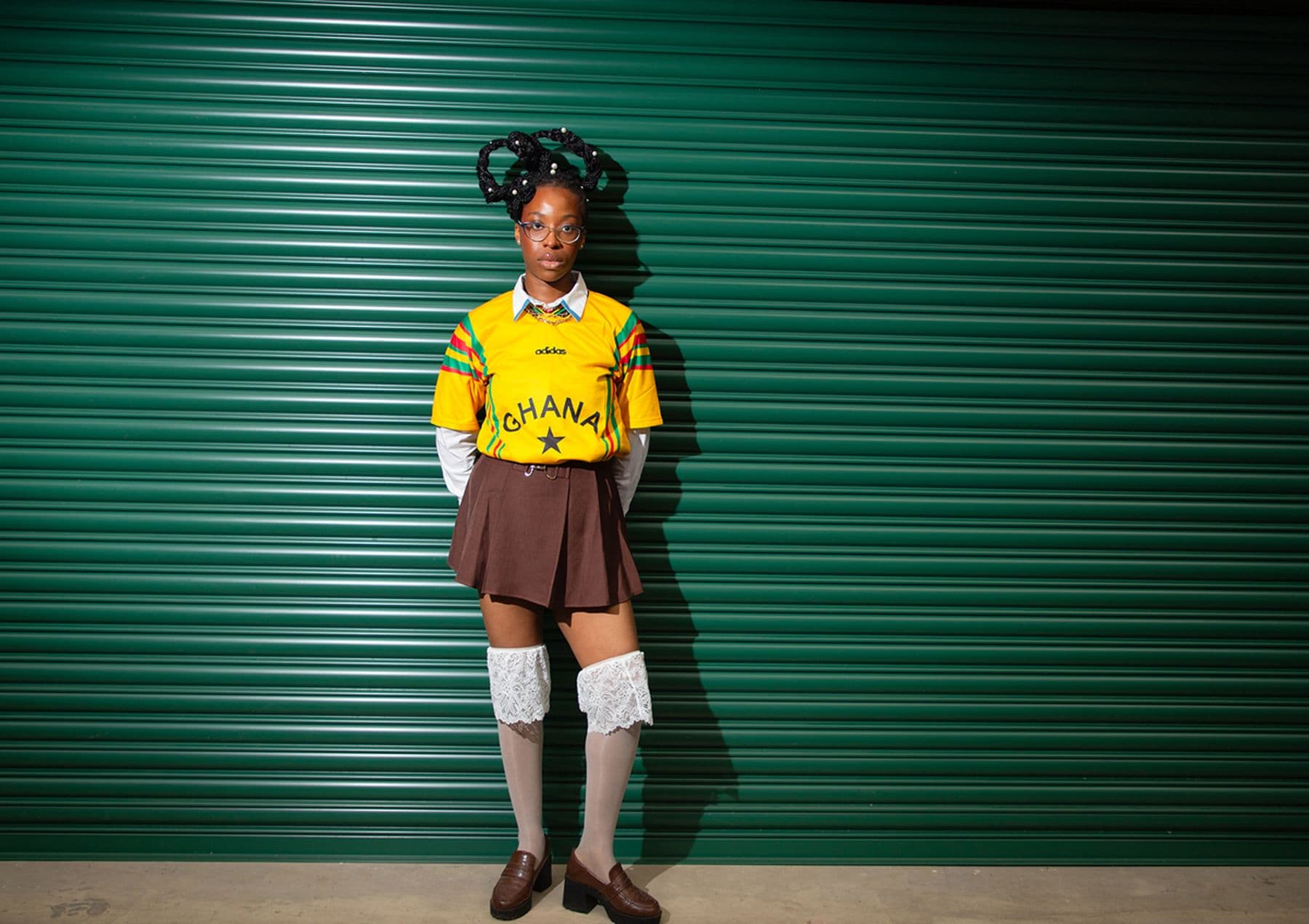 A photograph featuring a person in a Ghana soccer jersey and brown skirt standing against a green corrugated metal wall wearing knee-high socks and glasses.