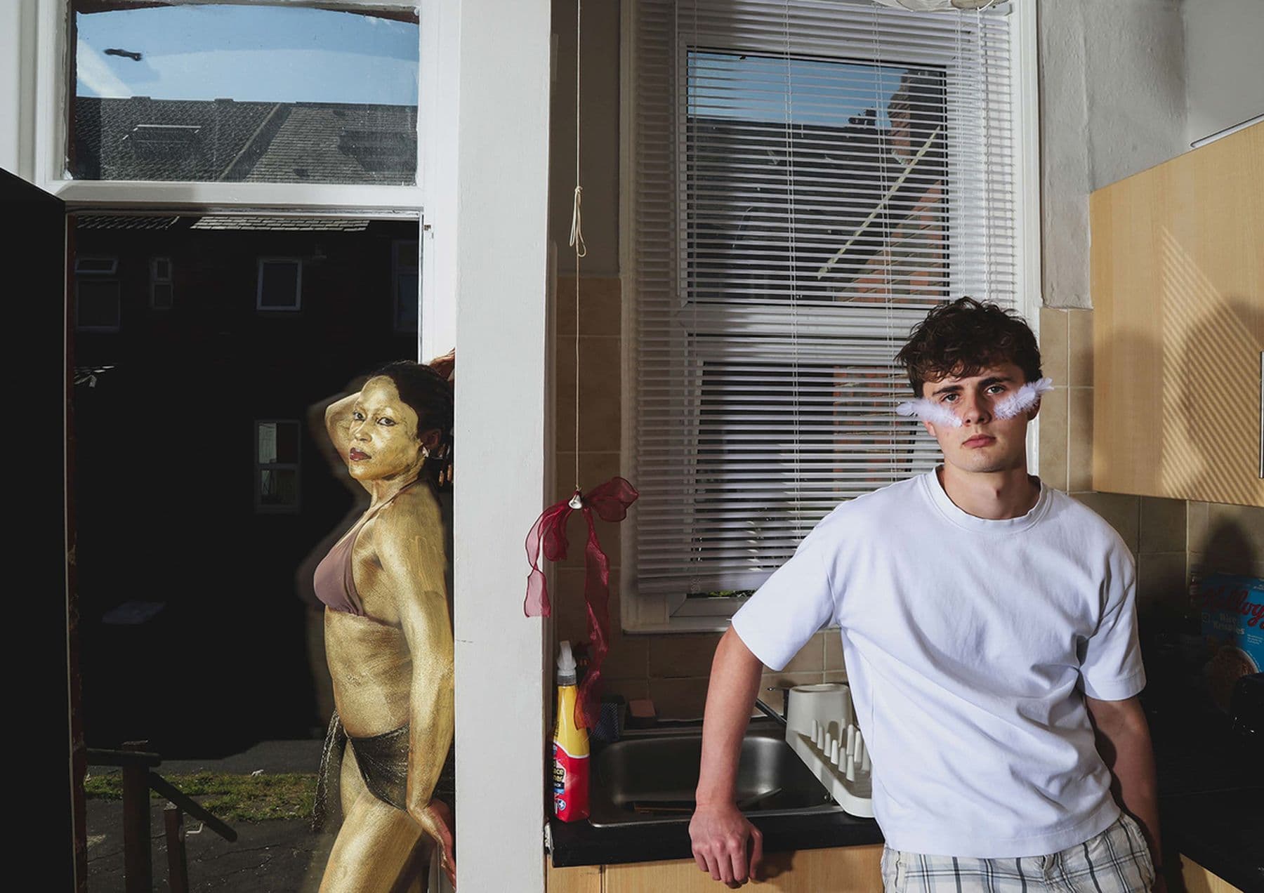 A photograph featuring a person posing in a white t-shirt while standing in a kitchen. In the hallway a person painted gold and wearing a bikini poses against the wall.