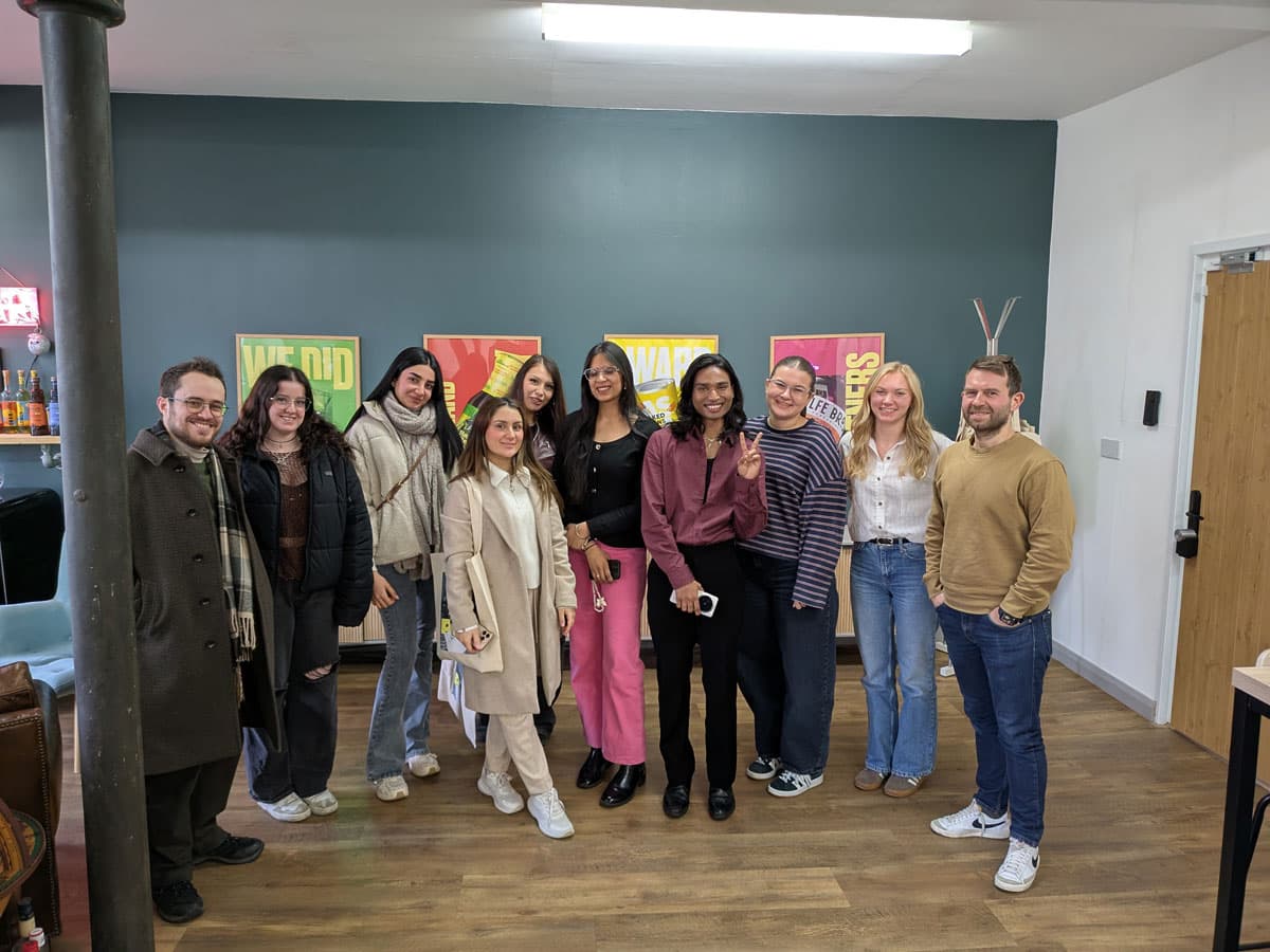 A group of nine people standing indoors, smiling at the camera. Framed motivational posters hang on the wall behind them.
