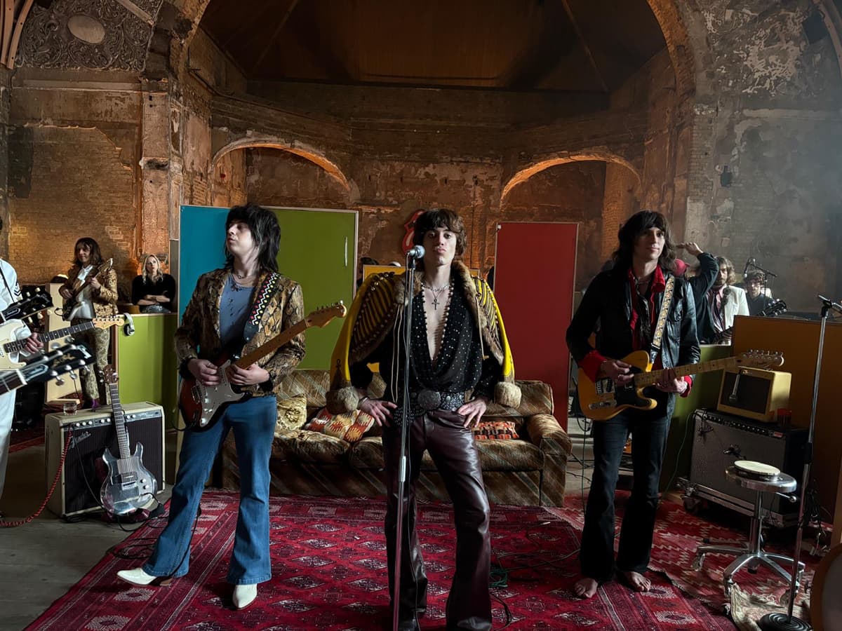 A vintage-style rock band performs in a dimly lit, rustic room with colourful walls and music equipment in the background.
