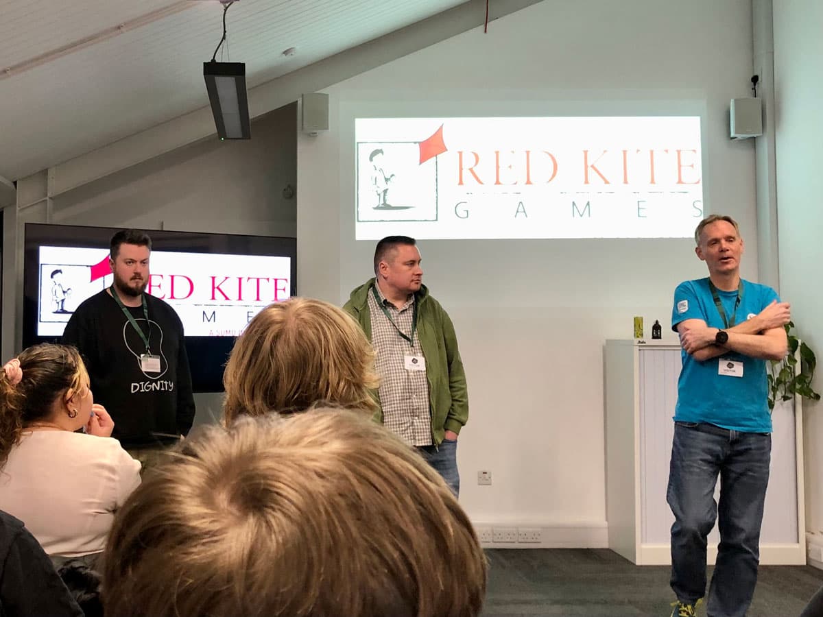 Three men stand in front of an audience at a Red Kite Games presentation. Two screens display the company logo.