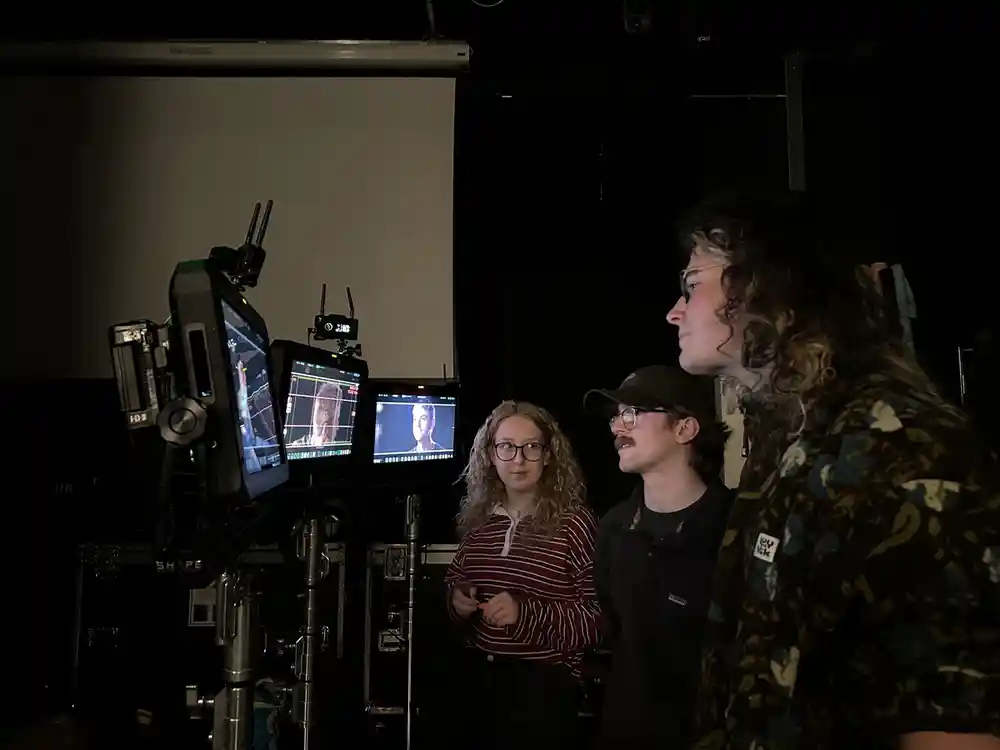 A photograph of three people standing in a dimly lit studio, observing a set of monitors displaying images, in a film or video production setting.