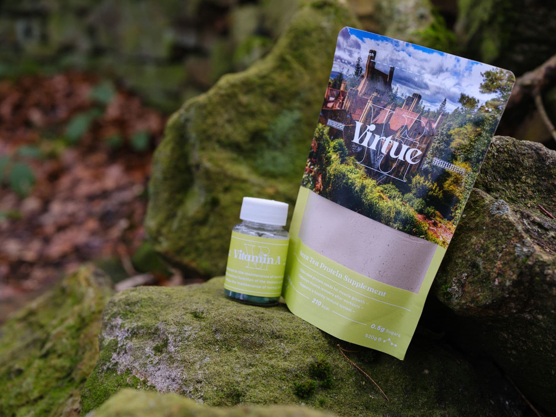 A branded packet and bottle are positioned on a mossy rock in a forest. The branding features a photo of a building and light lime green background with white text that reads: Virtue.