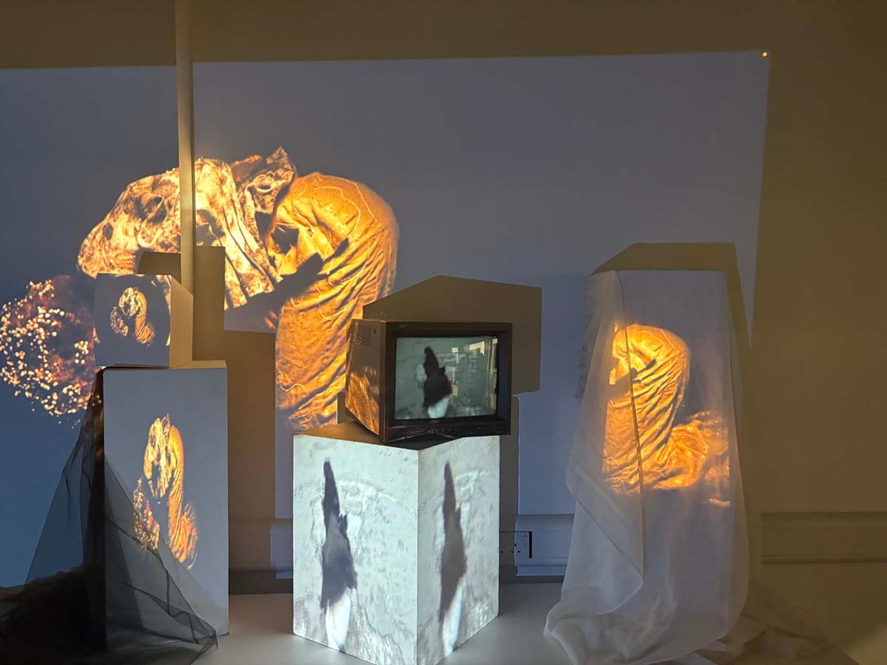 A photograph of an art installation featuring abstract projections on three draped objects and a screen creating a dynamic light and shadow effects.
