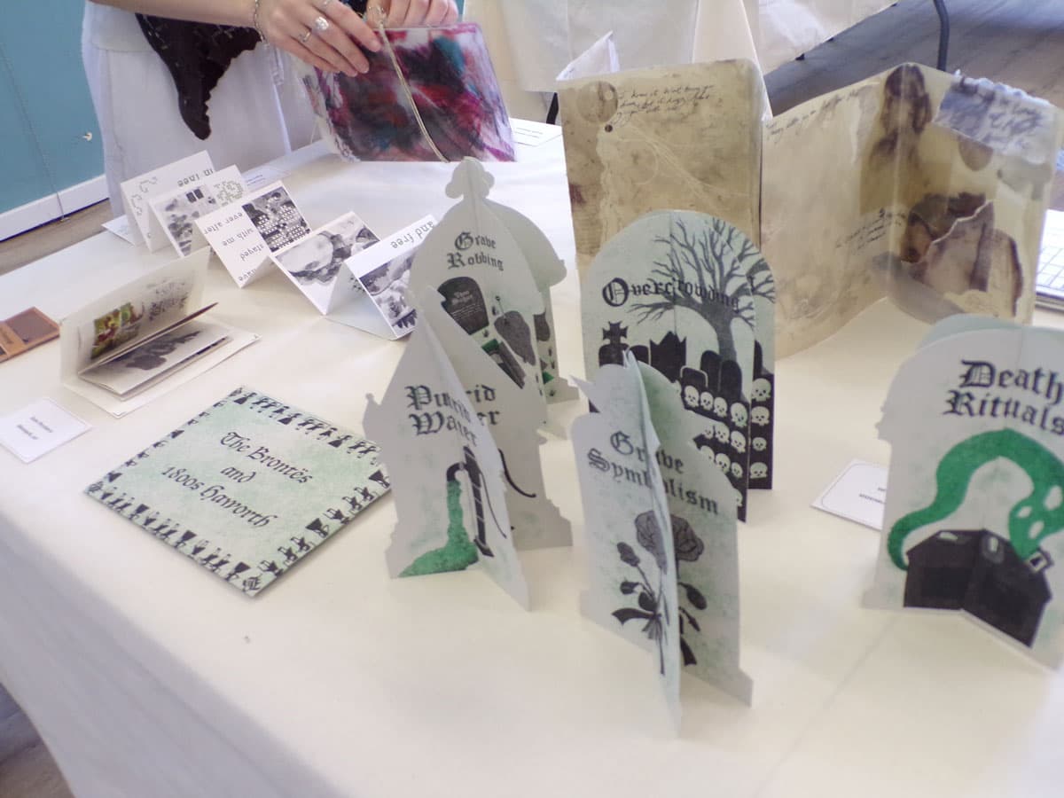 A table displays art books and pop-up cards exploring themes like death rituals and grave symbolism, with a person examining one item.