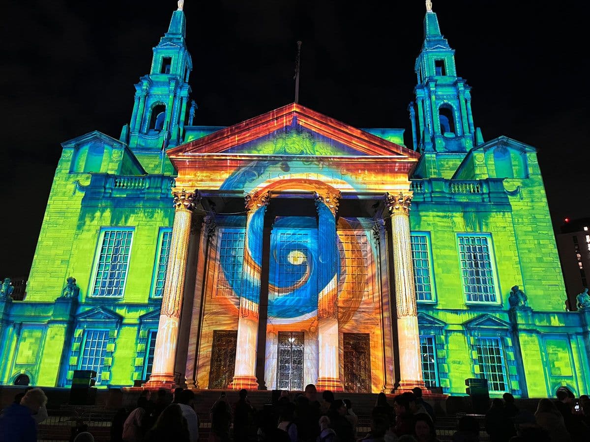 Illuminated building with colourful light projections, featuring swirling patterns on the facade, and two tall spires against a night sky.
