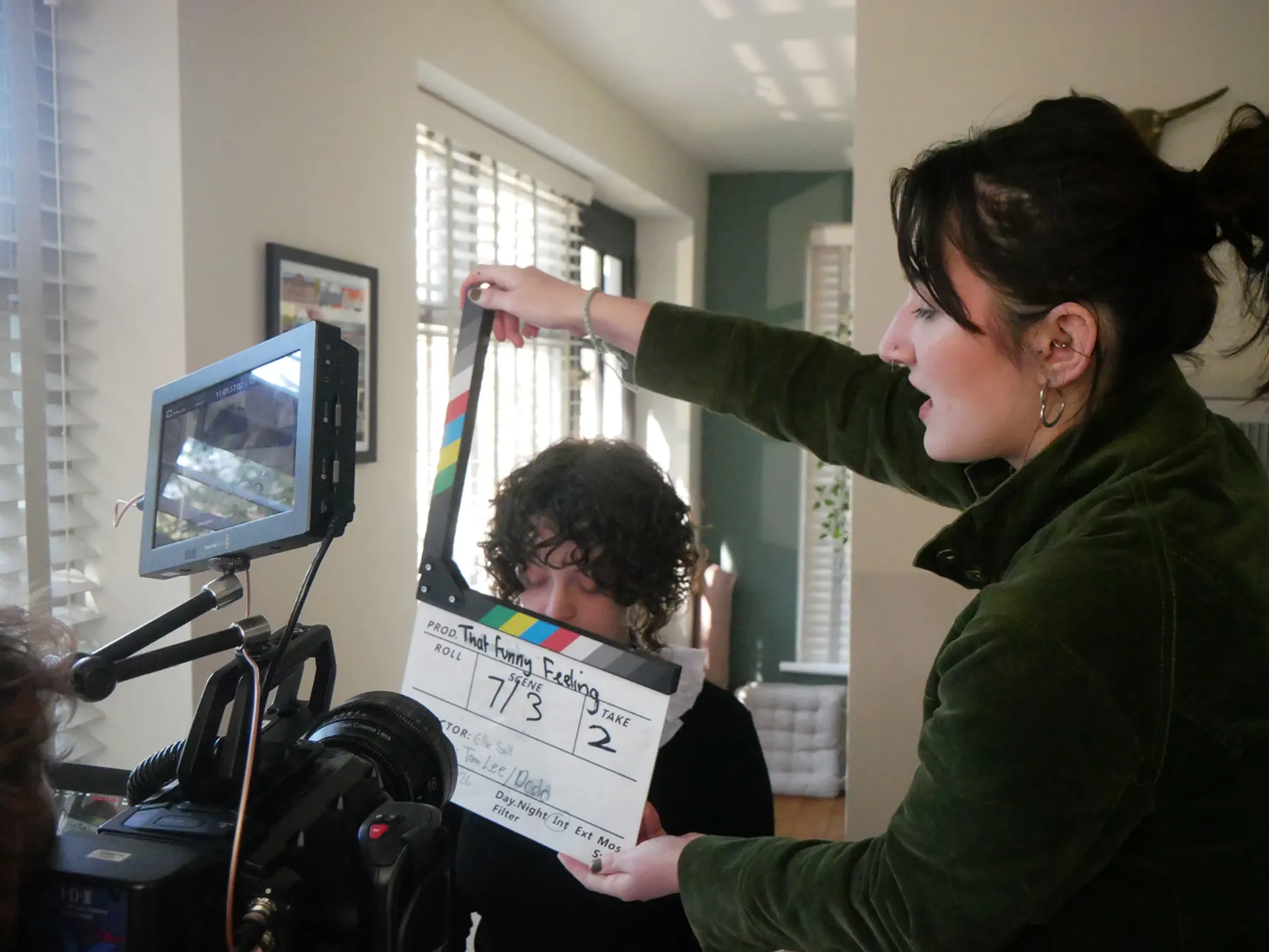 A person holds a clapperboard near a seated person on a film set with a camera and monitor showing a live feed in the foreground.