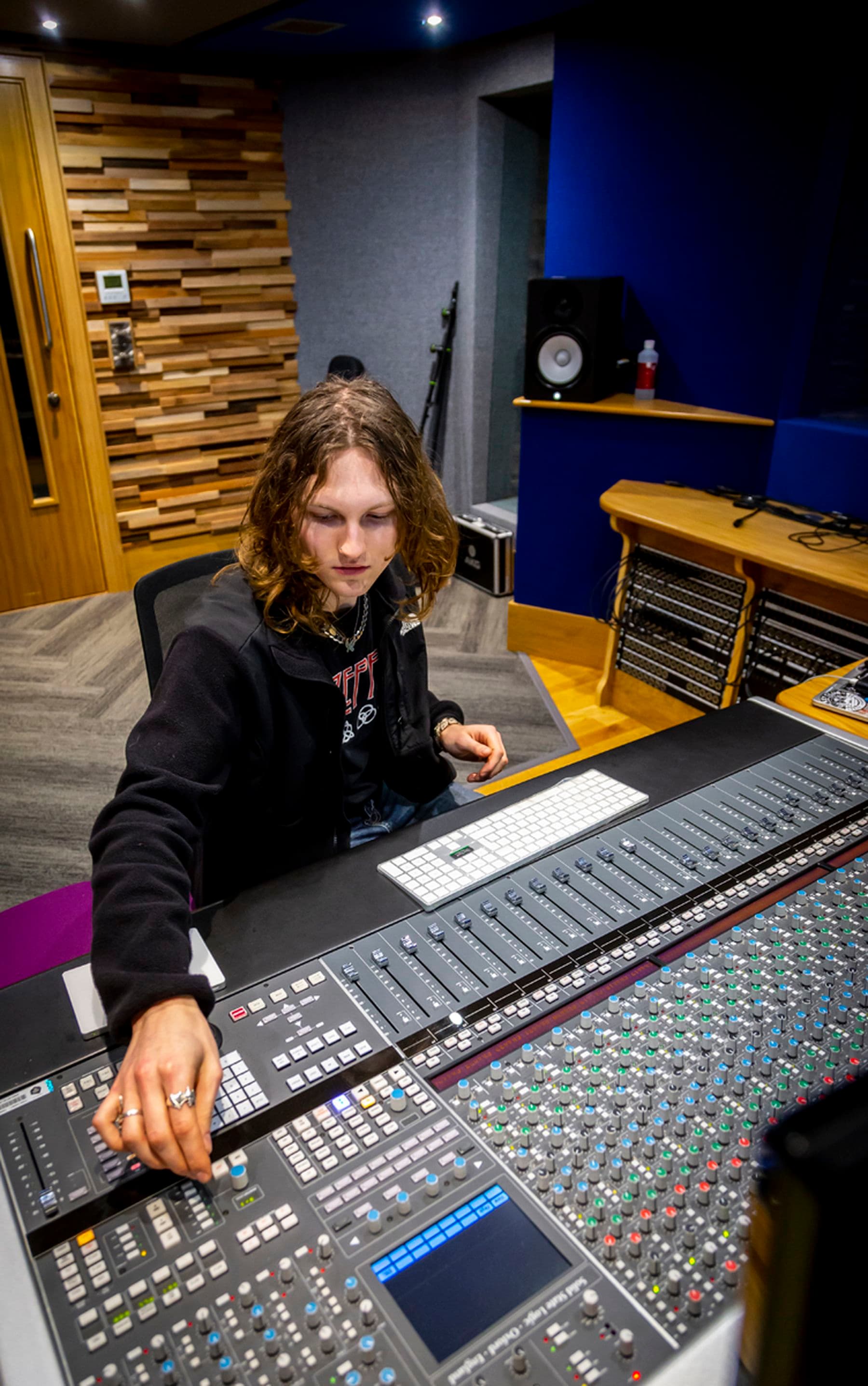 A photograph of a student sitting down and turning a dial on a large channel mixing console inside a recording studio.