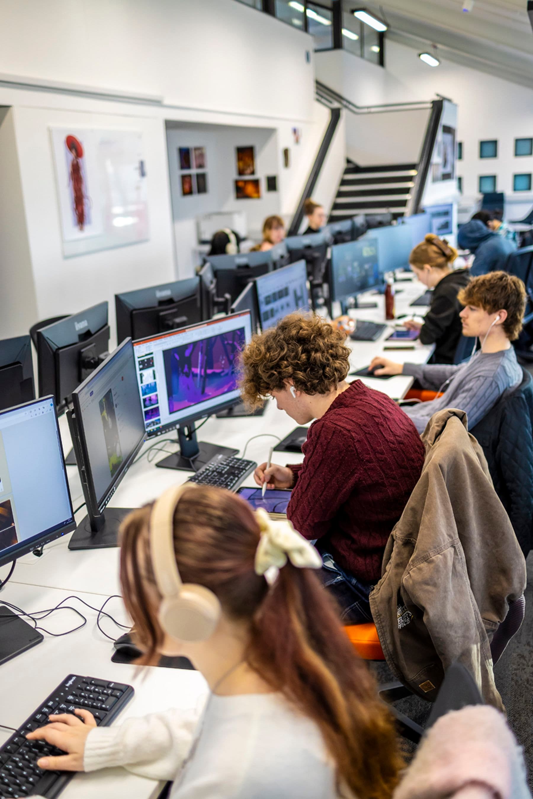 Students working on computers in a modern studio, focusing on digital design projects. Headphones and jackets are visible on chairs.