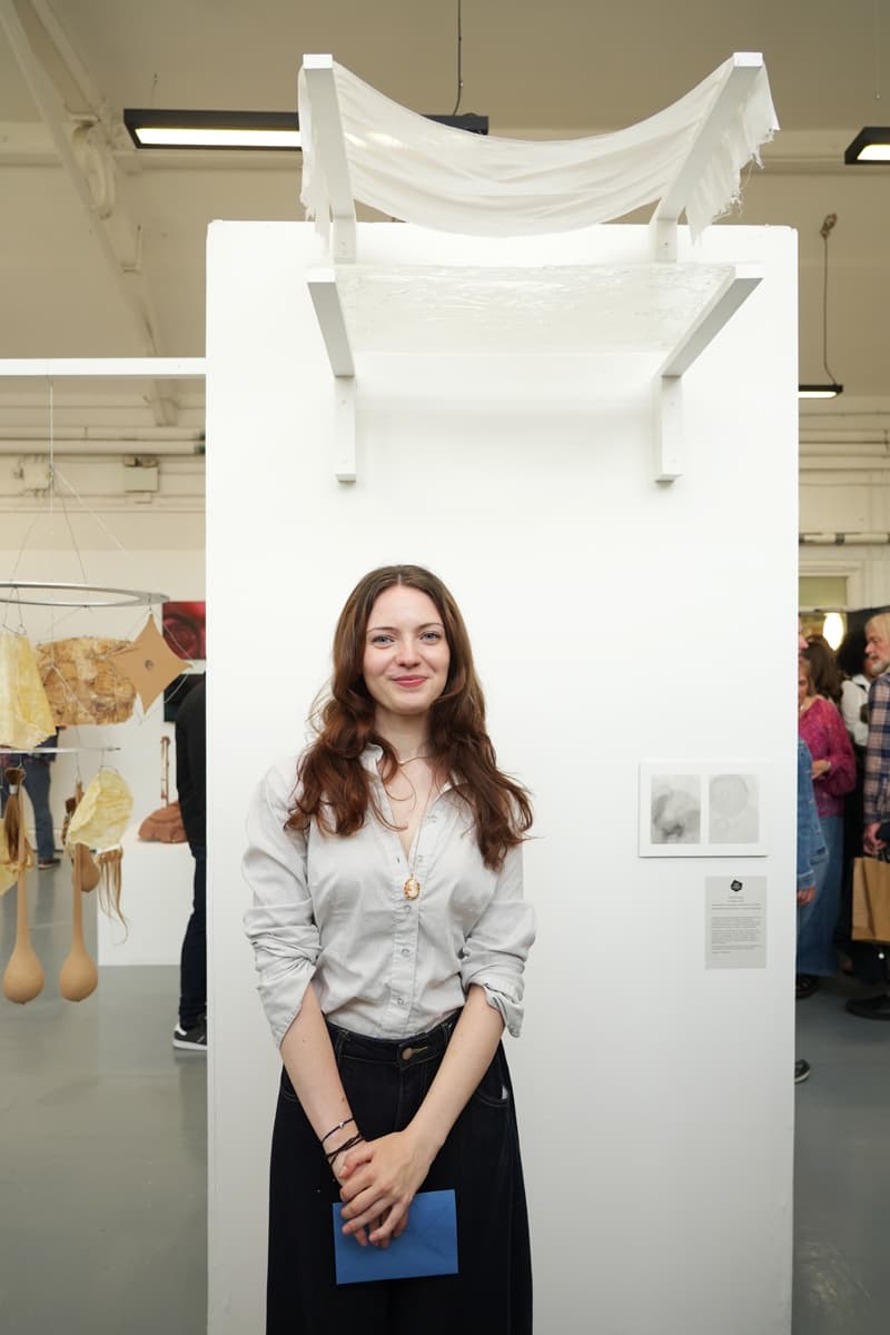 Person standing in front of white art display with fabric sculpture above. They are smiling and holding a blue envelope. Art pieces hang in the background.