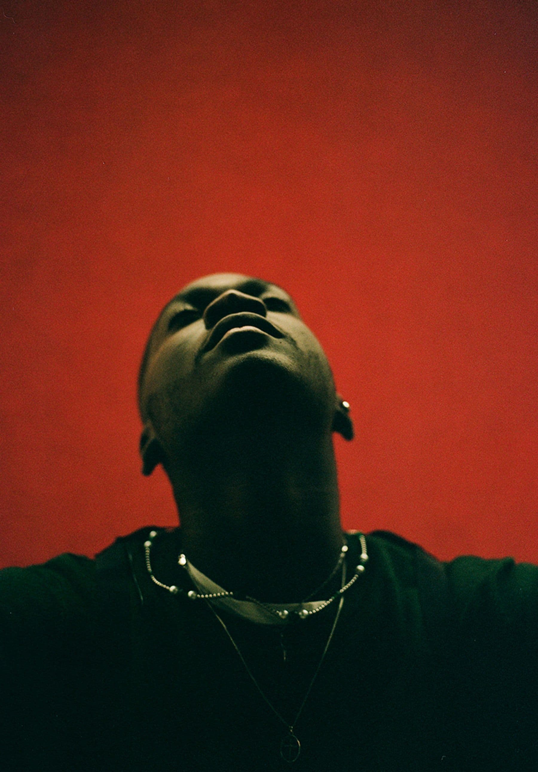 Person in a dark outfit with head tilted back against a red background, wearing layered necklaces.