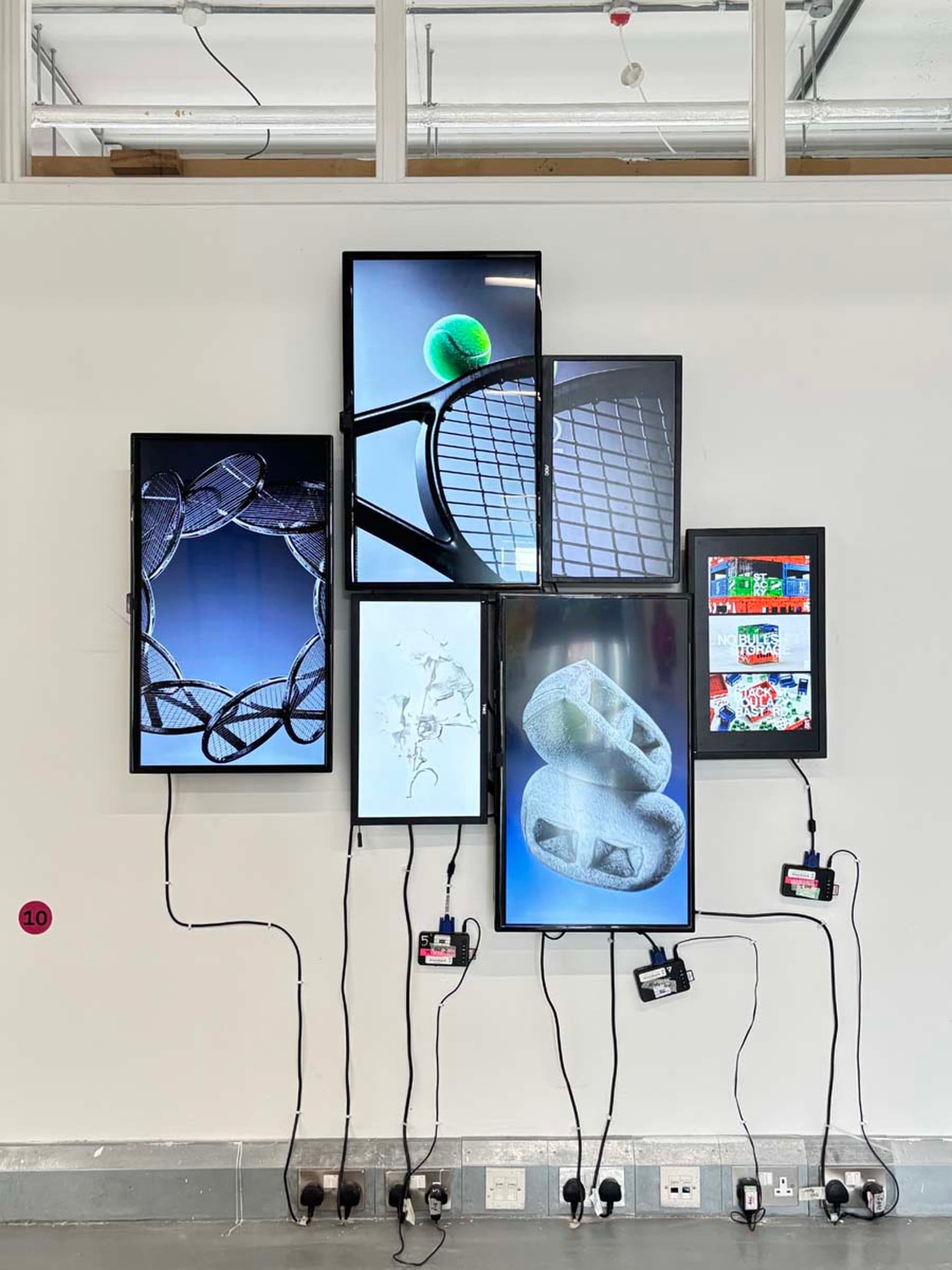 A photograph of a wall display of six digital screens featuring tennis-themed images, like rackets and balls, interconnected with visible cables and adapters below.
